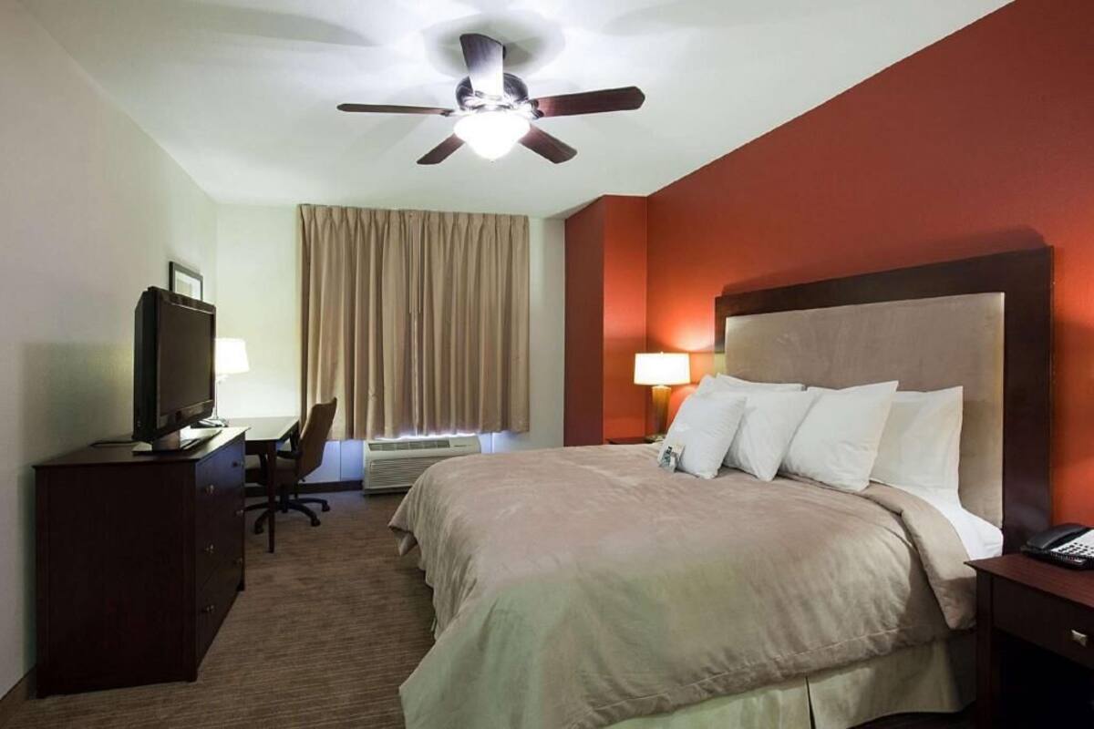 A hotel room with a bed, a desk with a television, a chair, a ceiling fan, and a window with curtains.