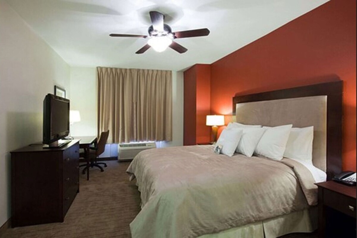 Habitación de hotel con cama, escritorio con computadora, televisión, ventilador de techo y ventana con cortinas.