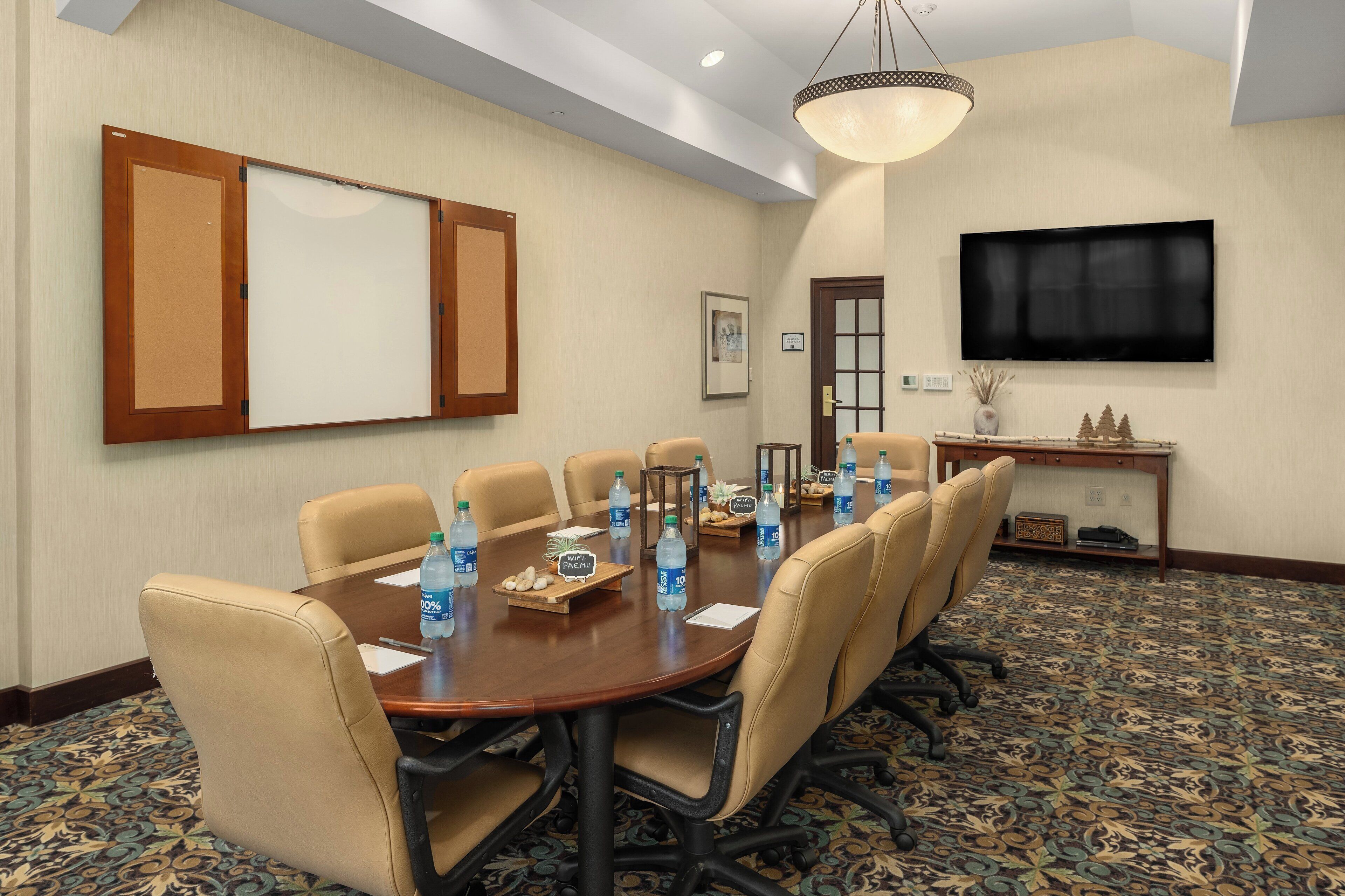 A conference room with a long table, beige chairs, water bottles, and a TV on the wall.