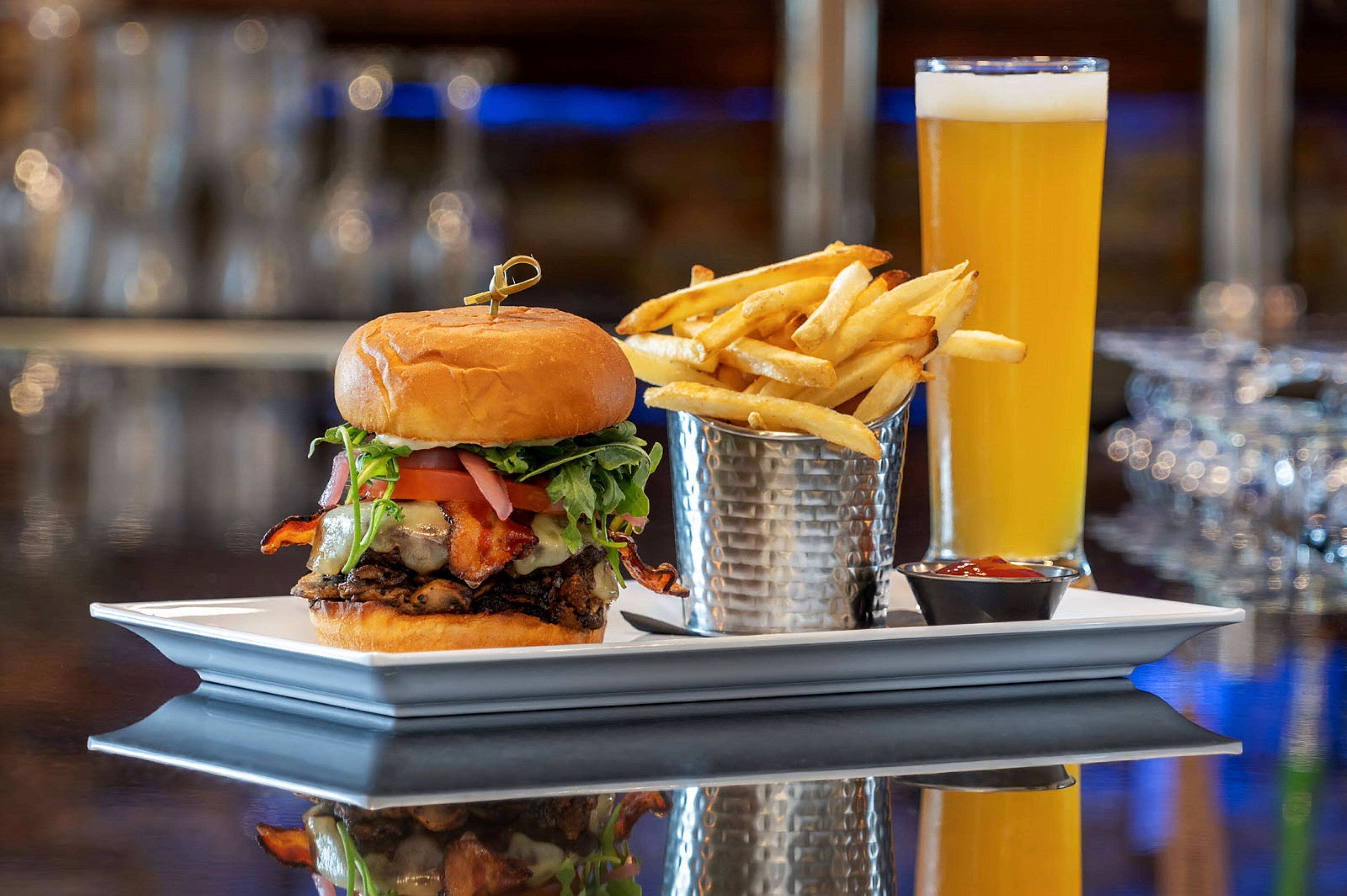 Hamburguesa con papas fritas y una cerveza sobre una superficie reflectante.