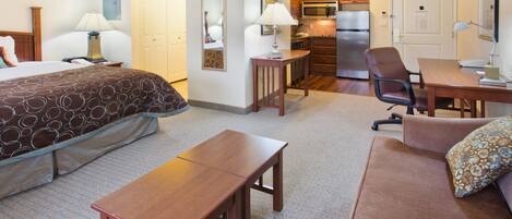 A hotel room with a bed, a wooden table, a sofa, a desk, a chair, a lamp, a mirror, a kitchen area with a refrigerator, microwave, and cabinets.