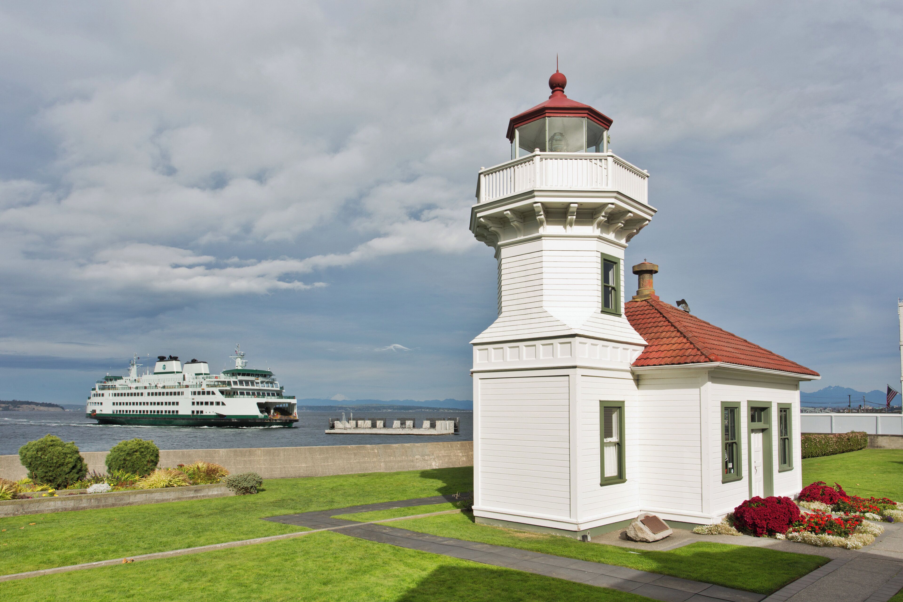 Un faro blanco con techo rojo, una zona verde con flores y un barco grande al fondo.
