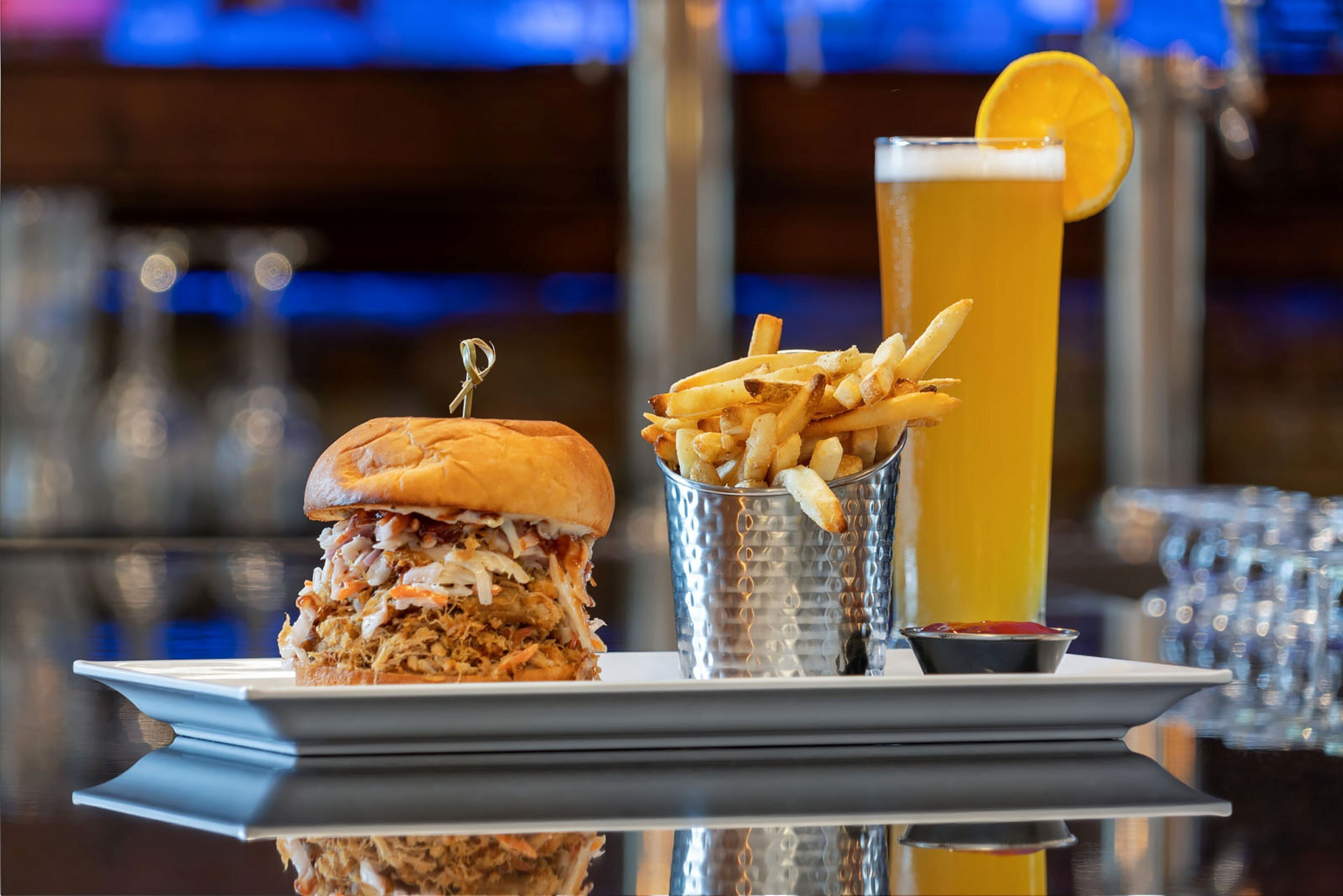 Un plato que incluye un sándwich de cerdo desmenuzado, papas fritas y un vaso alto de cerveza con una rodaja de limón.