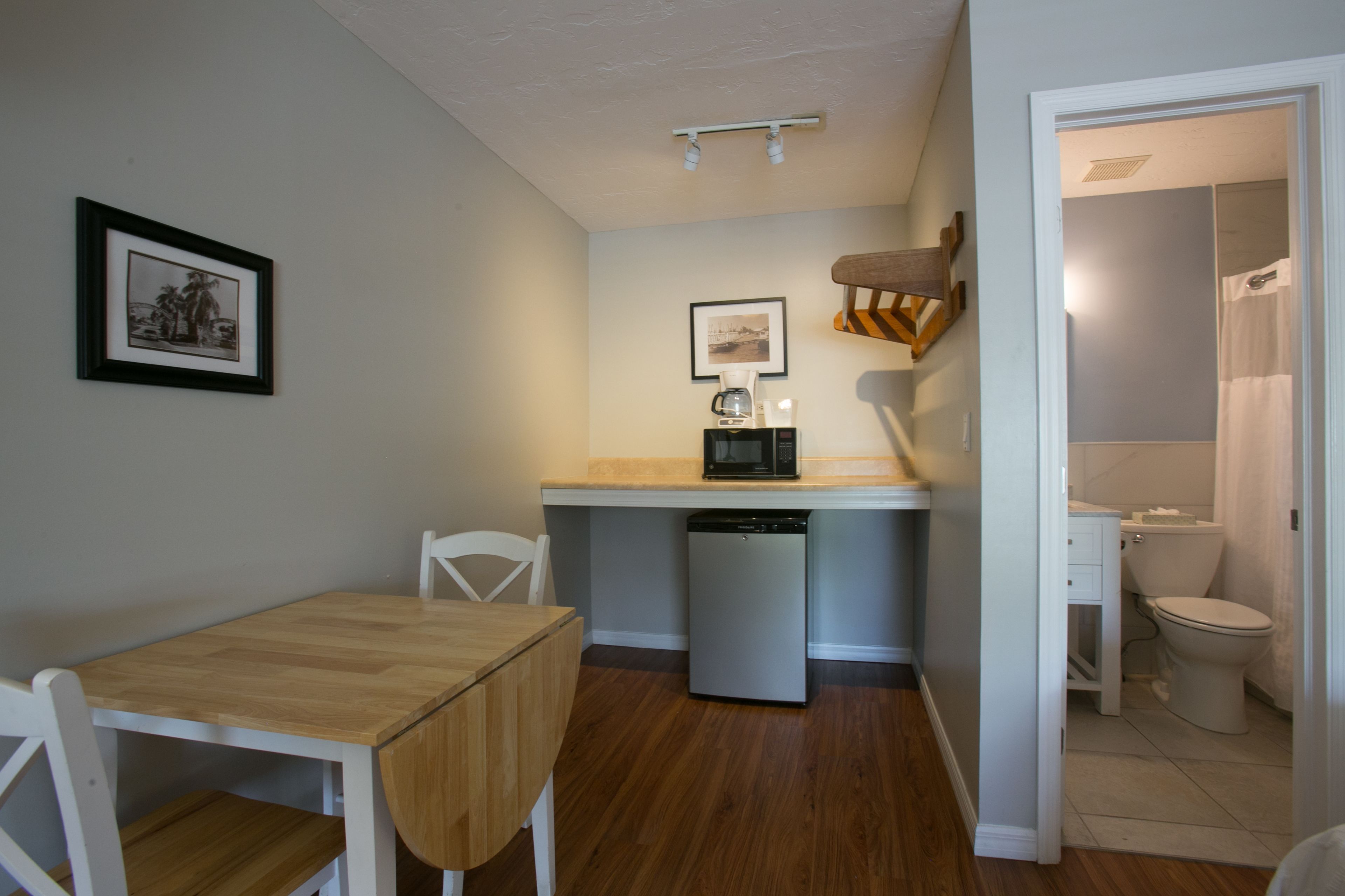 A compact kitchen with a microwave, coffee maker, and a small refrigerator. There is a dining table with chairs and a framed picture on the wall.