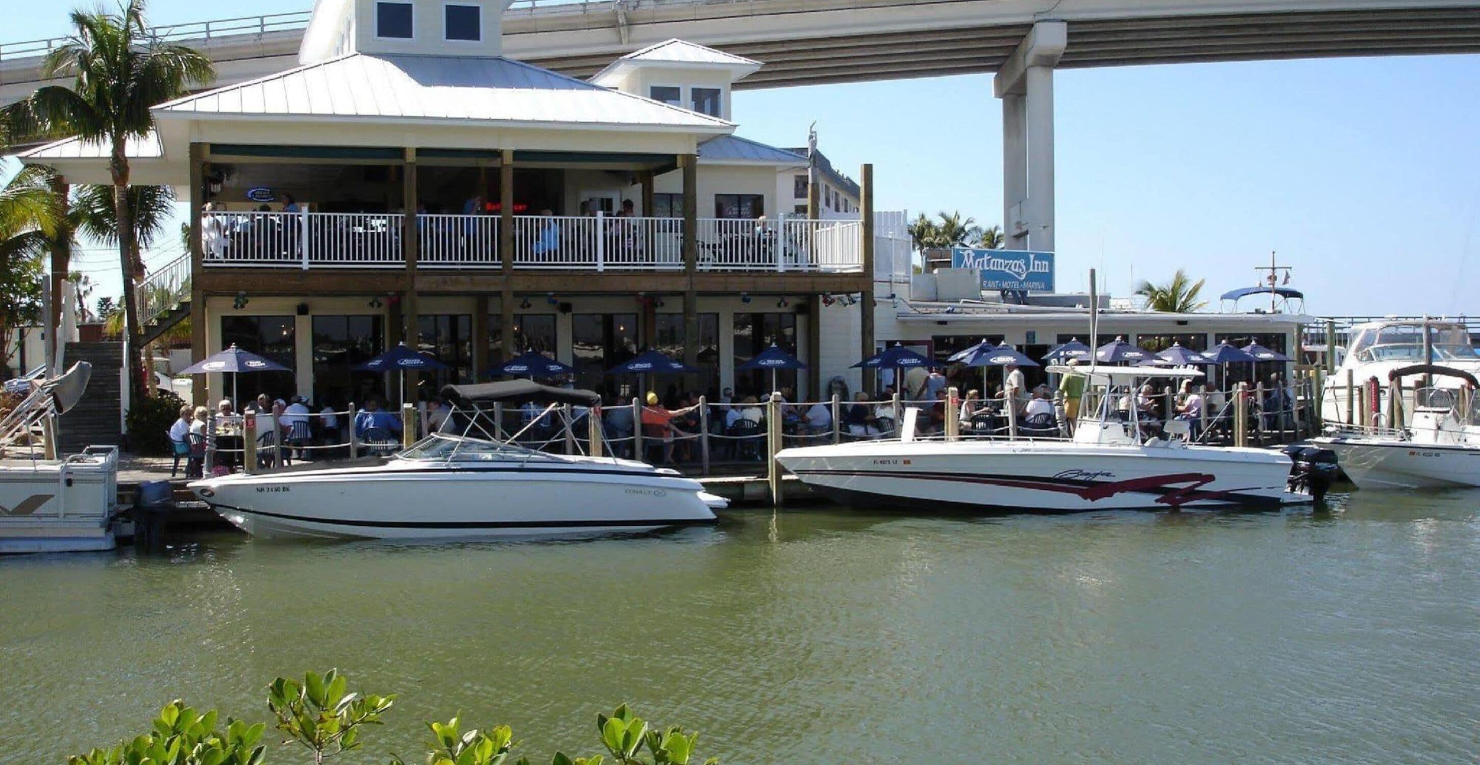 A marina with boats docked, a restaurant with outdoor seating, and a building with a balcony overlooking the water.