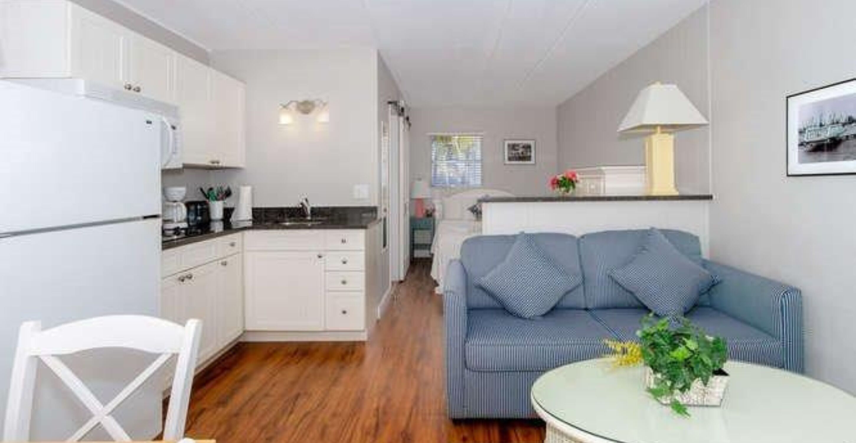 Un espacio habitable compacto con kitchenette, una zona de comedor con una silla blanca, un sofá con cojines azules y una mesa redonda con una planta.