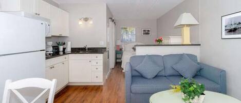 A compact living space with a kitchenette, a dining area with a white chair, a sofa with blue cushions, and a round table with a plant.