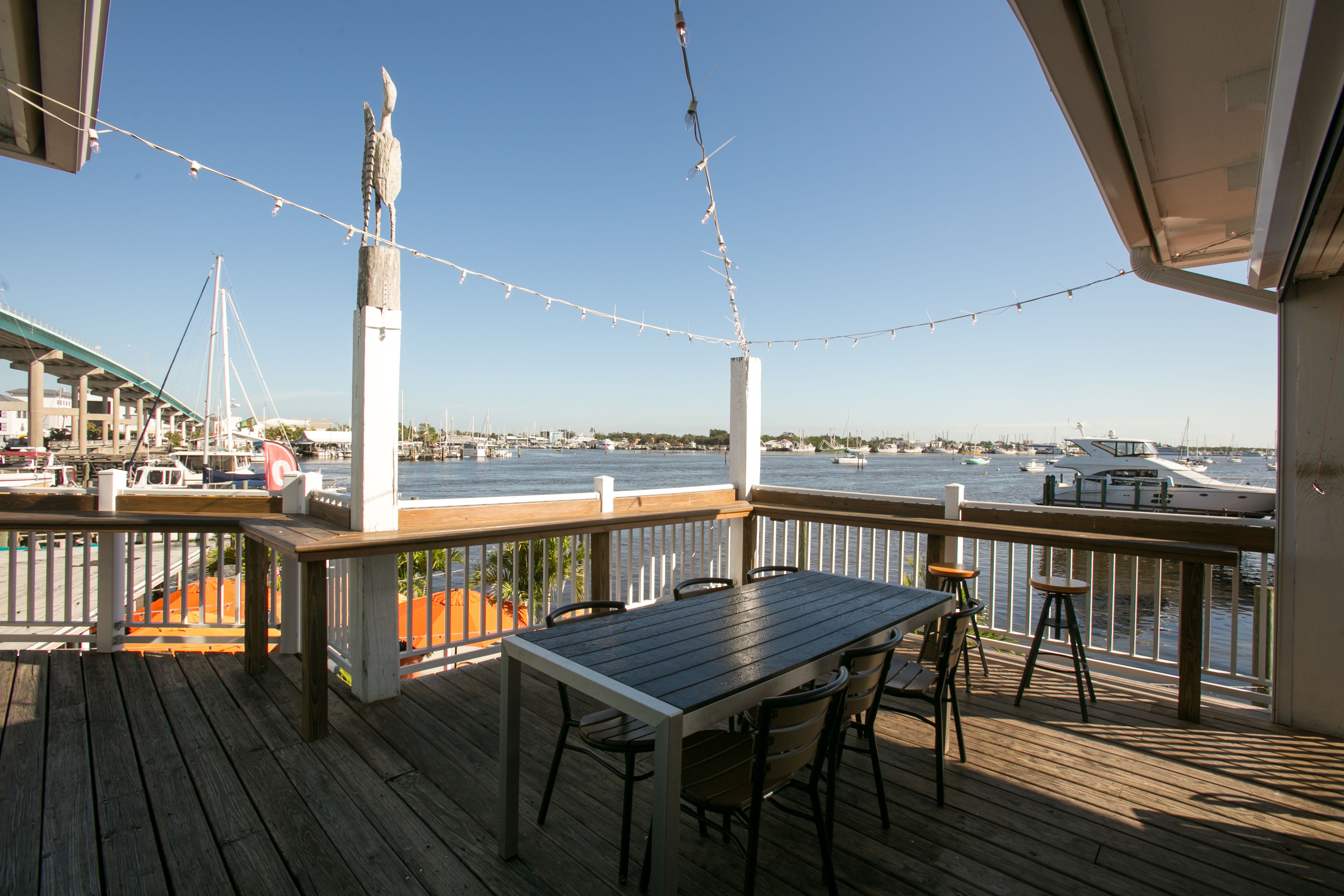A wooden deck with a table and chairs, overlooking a marina with boats and a clear sky.