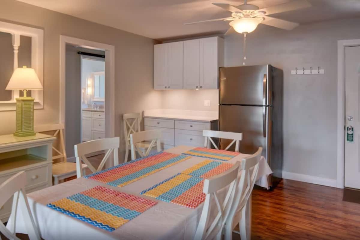 Una cocina con armarios blancos, un refrigerador de acero inoxidable y una mesa con un mantel individual colorido.