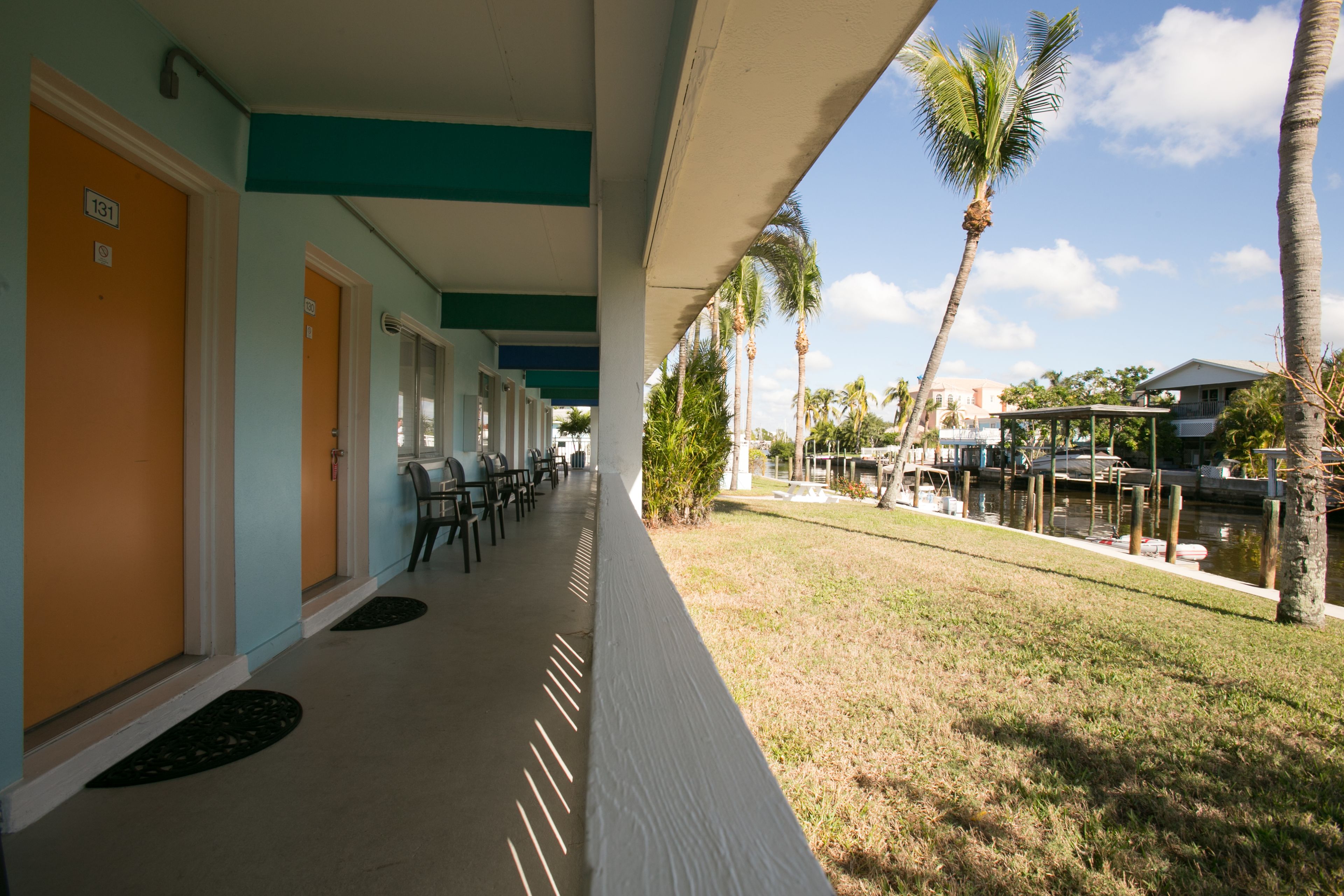 A hotel corridor with doors numbered 131 and 132, outdoor seating, palm trees, and a view of a dock with boats.