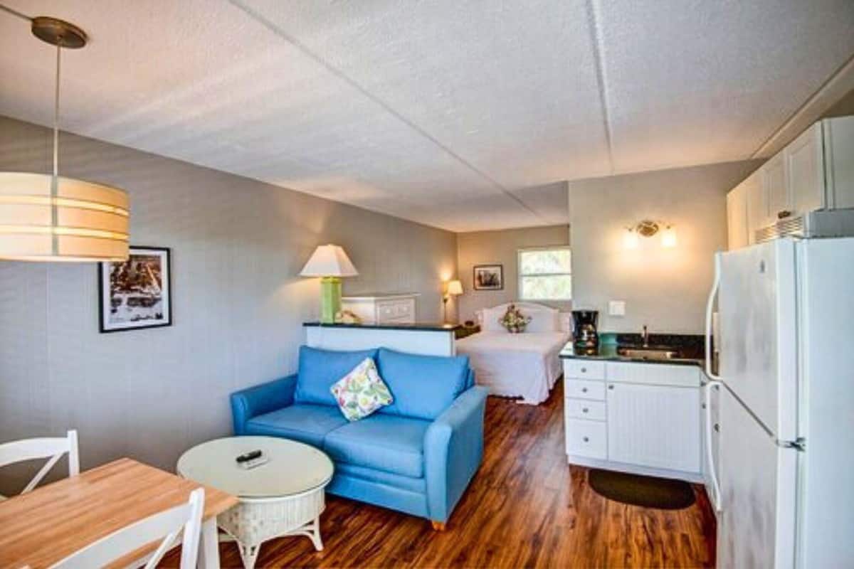 Habitación compacta con sofá azul, una mesita redonda pequeña, un armario blanco y una zona de kitchenette.