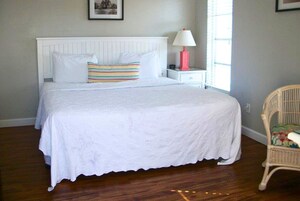 A neatly made bed with white linens and a striped pillow, a bedside table with a lamp, and a wicker chair.