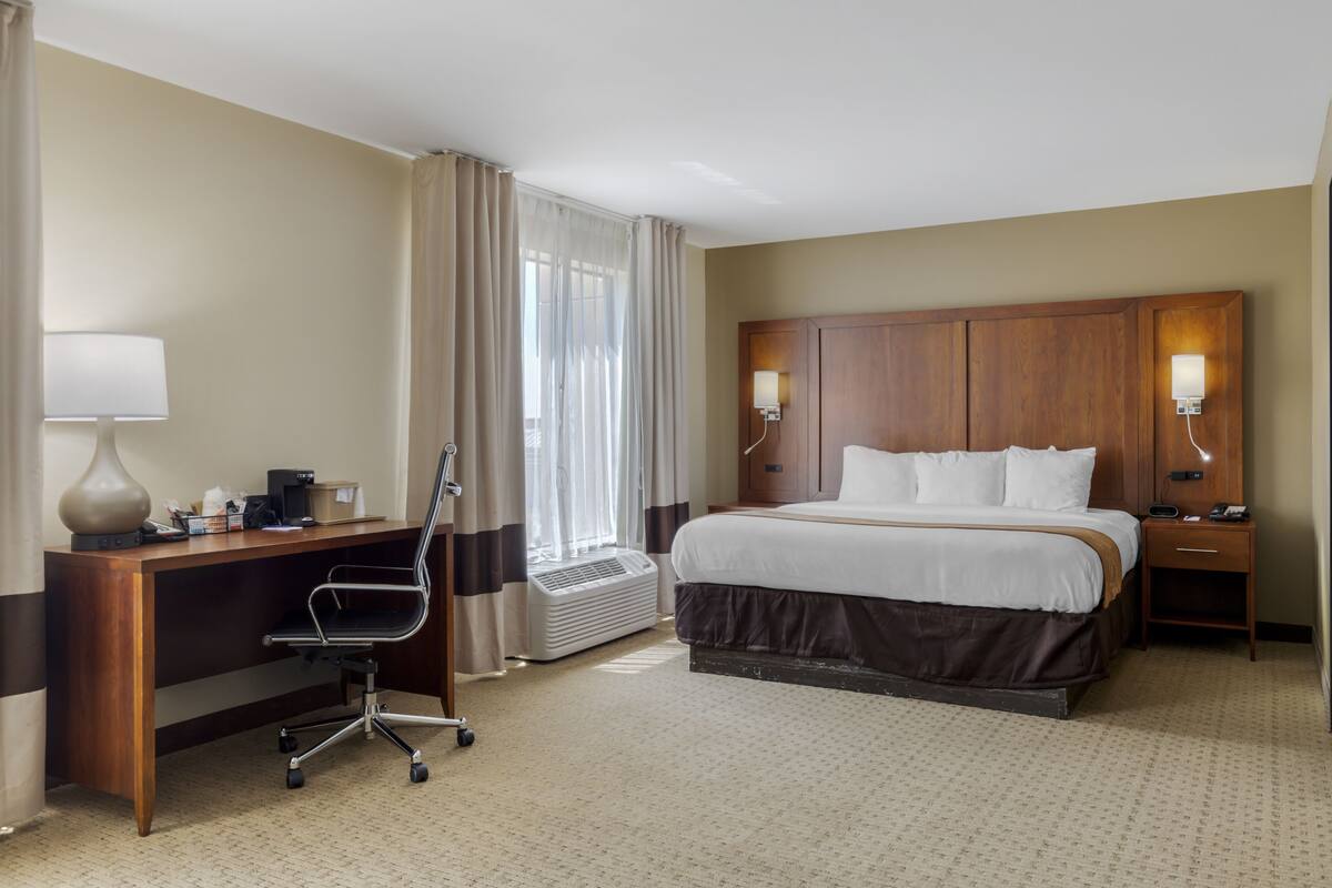 A hotel room with a bed, desk, chair, lamp, bedside table, and a window with curtains.