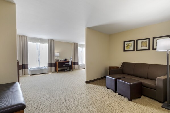 A hotel room with a large sofa, a desk with a chair, a lamp, and a window with curtains. - Comfort Suites Pflugerville - Austin North (Pflugerville)