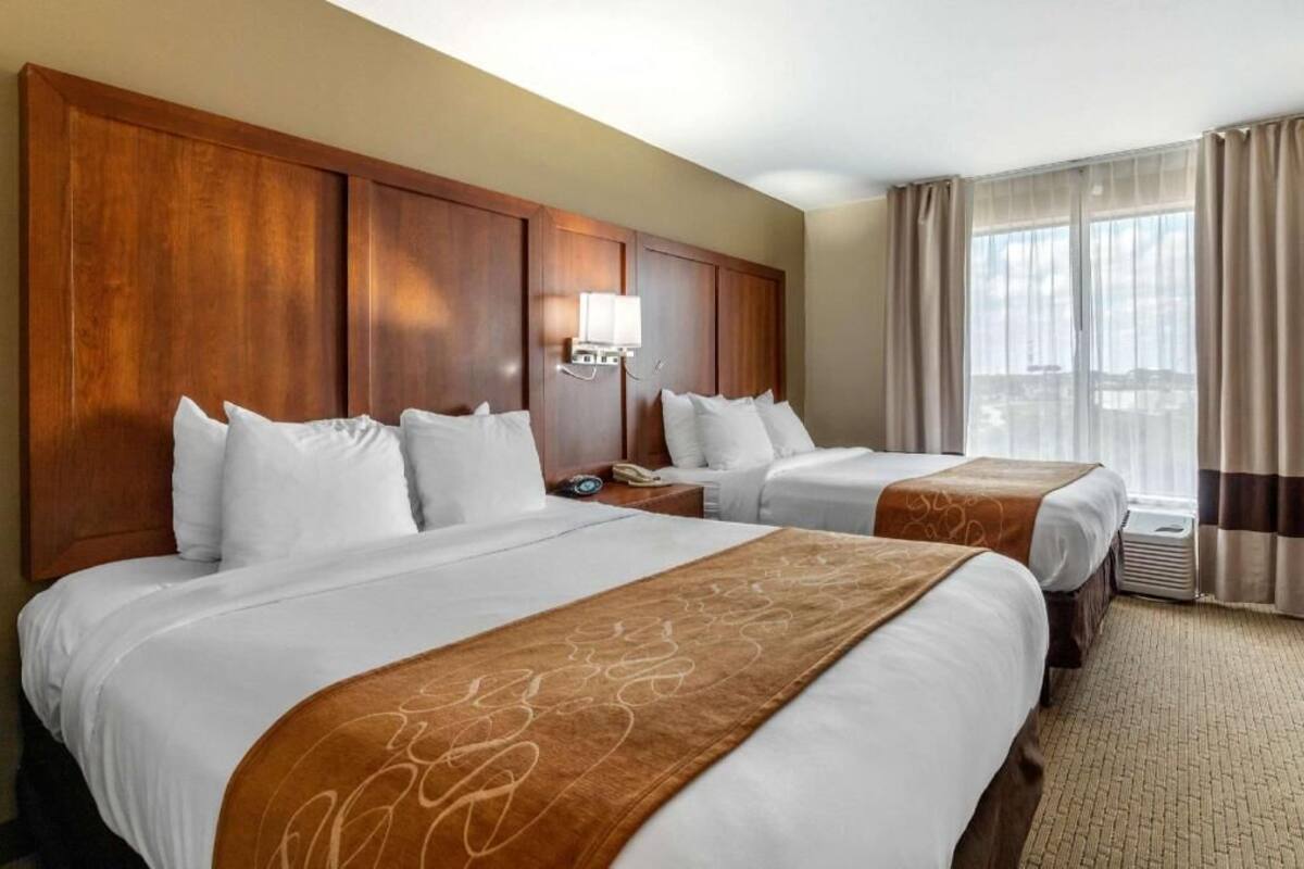 A hotel room with two beds, a wooden headboard, a nightstand with a lamp, and a large window with curtains.