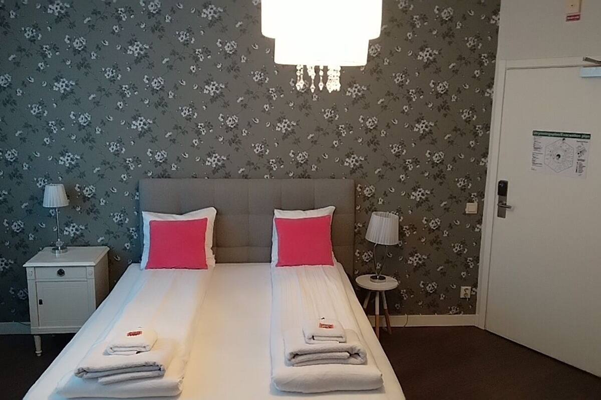 A hotel room with a bed, two pillows, a nightstand, a lamp, a door, and a wall with floral wallpaper.