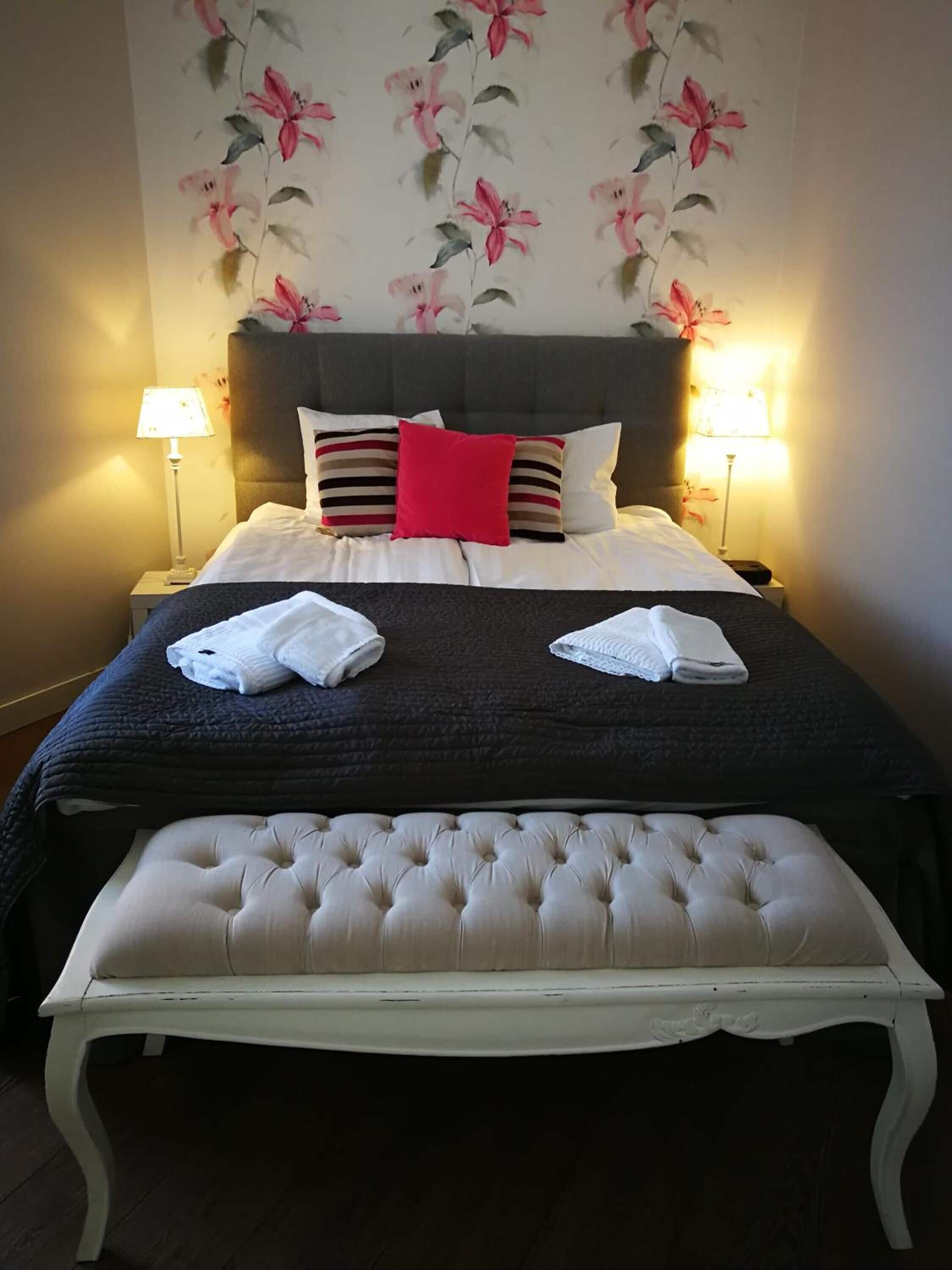 A neatly made bed with a dark headboard, two bedside lamps, and a floral wallpaper.