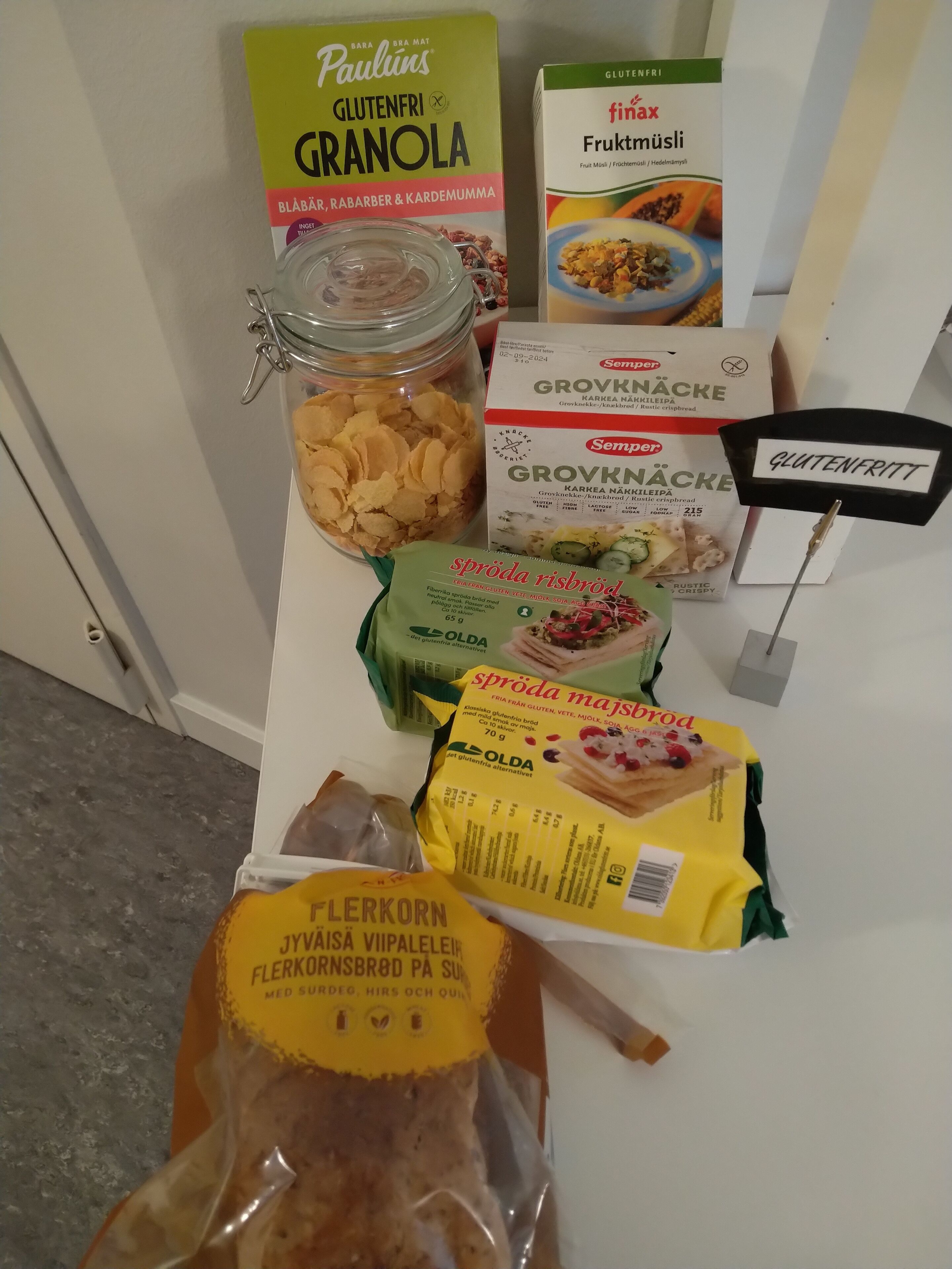 Variedad de productos sin gluten sobre una mesa, incluyendo granola, muesli de frutas, pan de semillas y un paquete de harina.