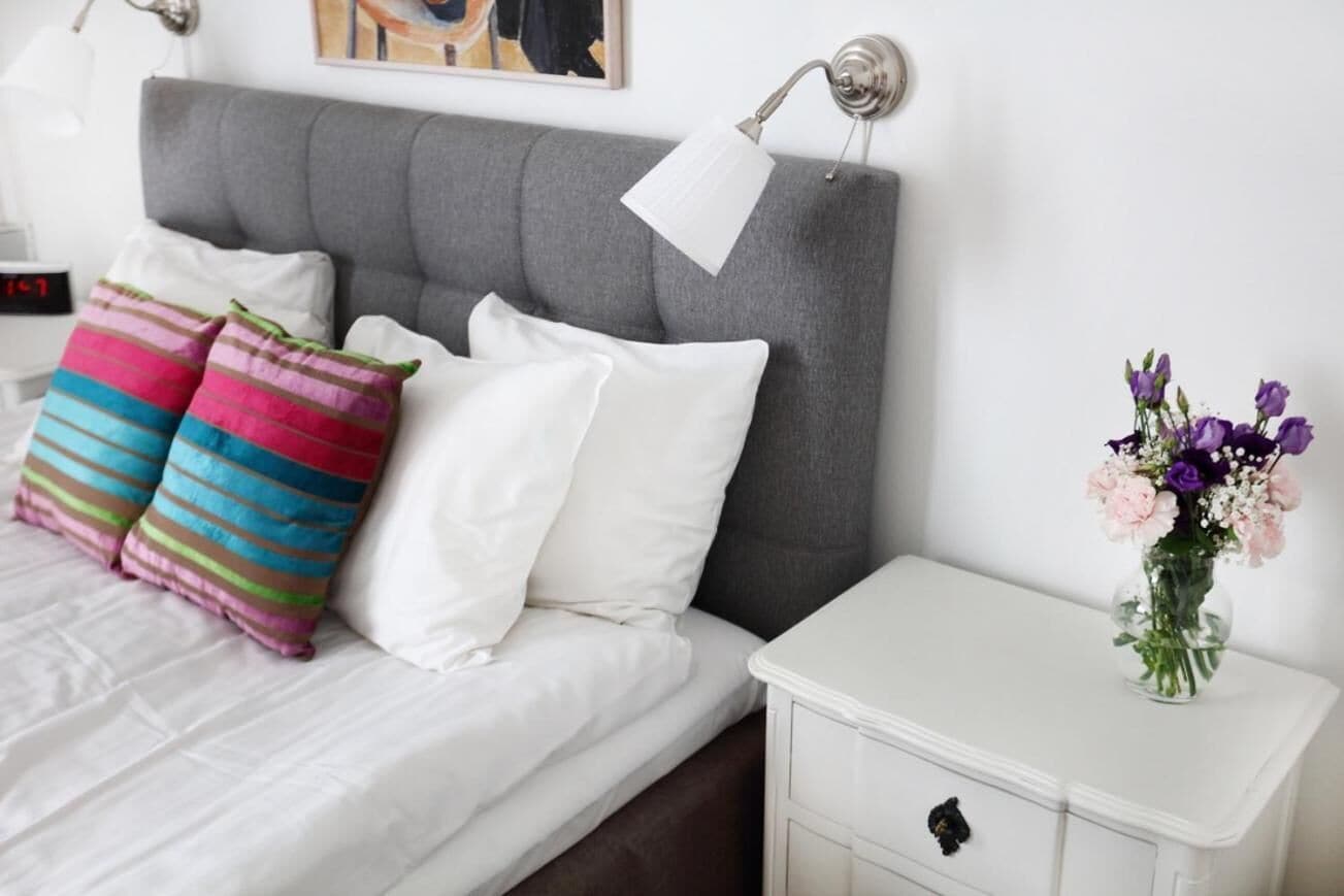 Una cama bien hecha con almohadas blancas y a rayas, un cabecero gris, una mesita de noche blanca con un jarrón de flores y un reloj en la mesita de noche.