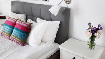 Una cama bien hecha con almohadas blancas y a rayas, un cabecero gris, una mesita de noche blanca con un jarrón de flores y un reloj en la mesita de noche.