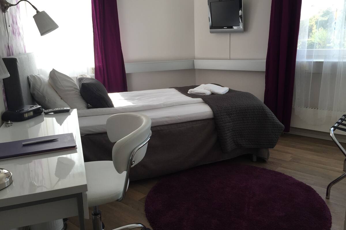 A hotel room with a bed, a desk with a chair, a TV mounted on the wall, and a window with curtains.
