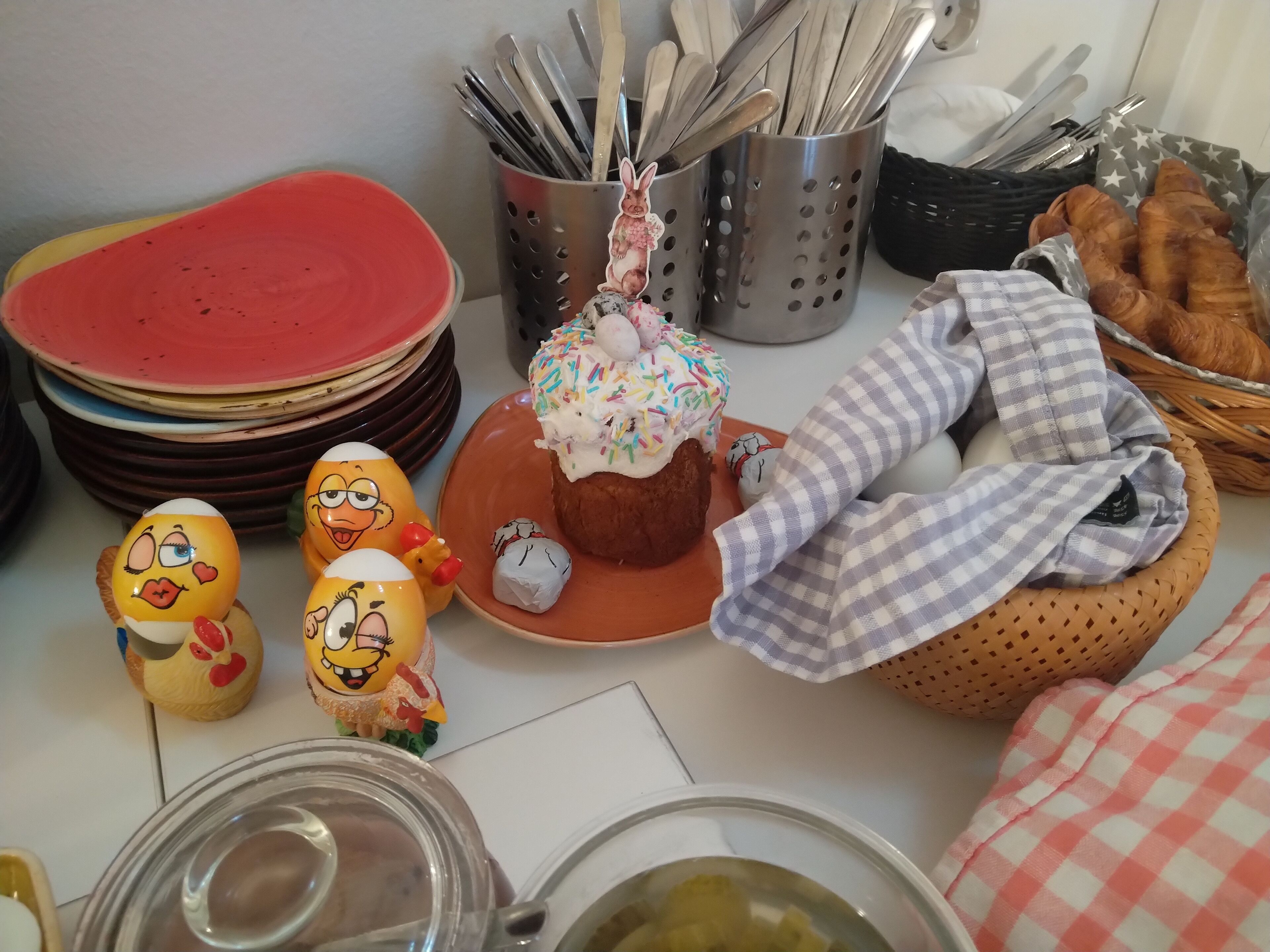 A table with a stack of plates, a basket of croissants, a plate with a decorated cupcake, and two figurines.