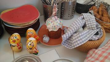 A table with a stack of plates, a basket of croissants, a plate with a decorated cupcake, and two figurines.