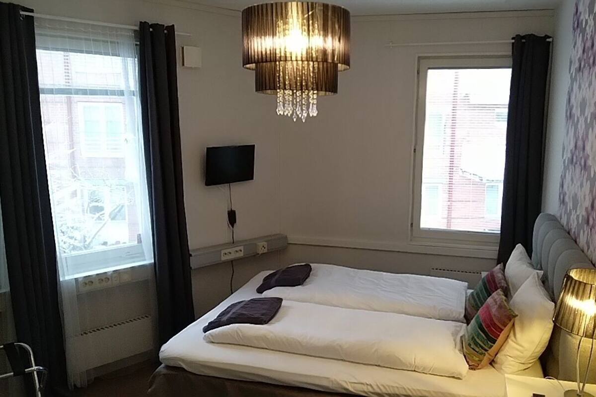 A hotel room with two beds, a TV mounted on the wall, a chandelier, and a window with curtains.