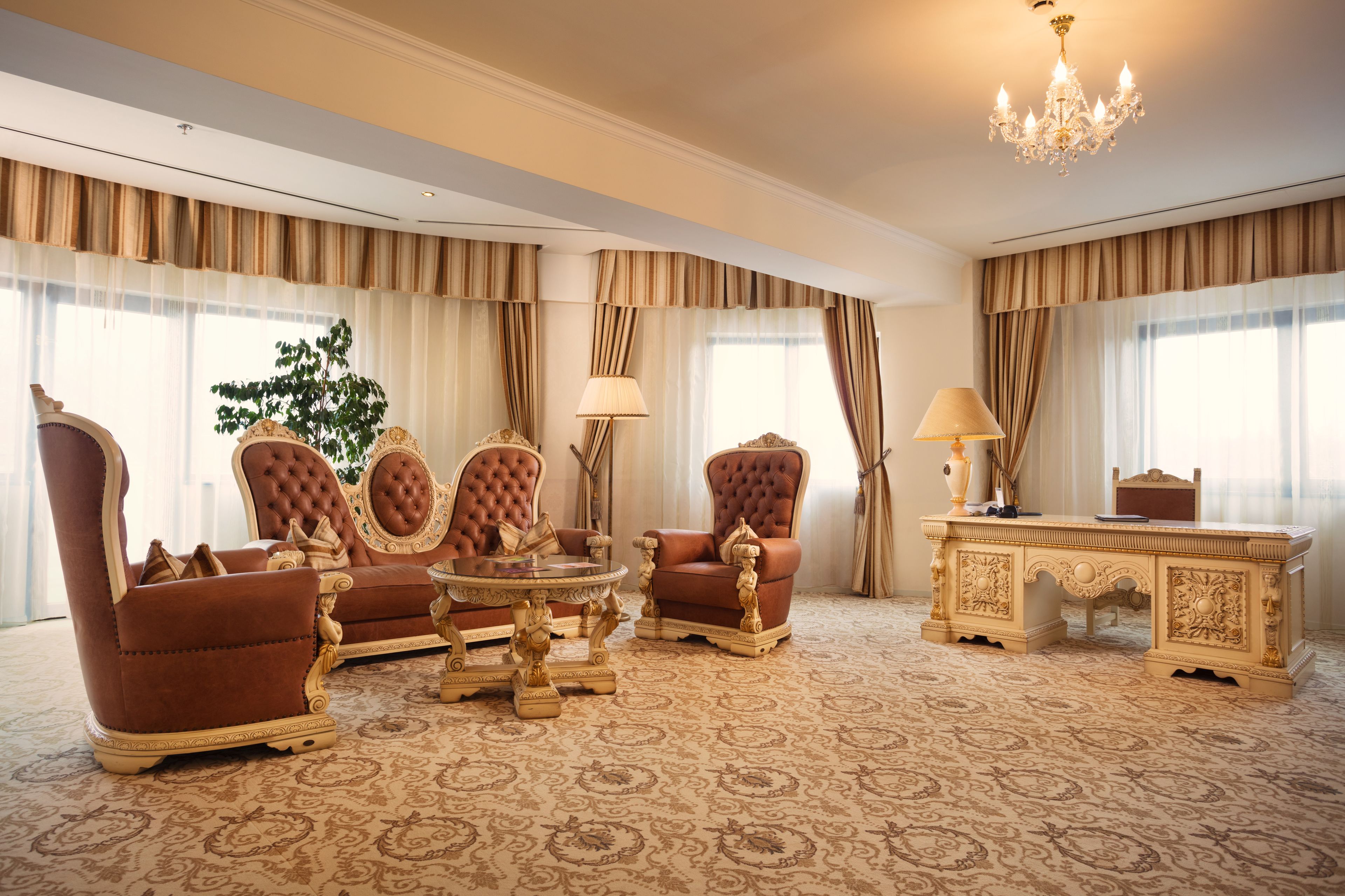 A luxurious room with a large, ornate desk, two brown leather armchairs, a round table, and a chandelier.