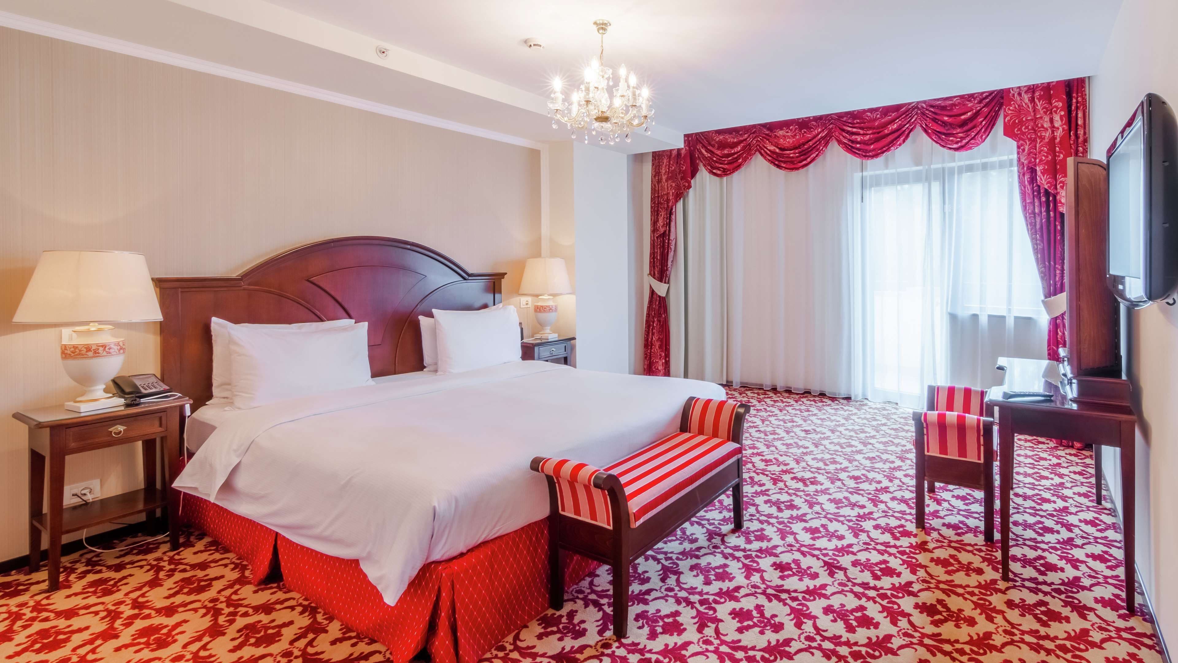A hotel room with a large bed, a bench, a desk with a chair, a TV, and a chandelier.