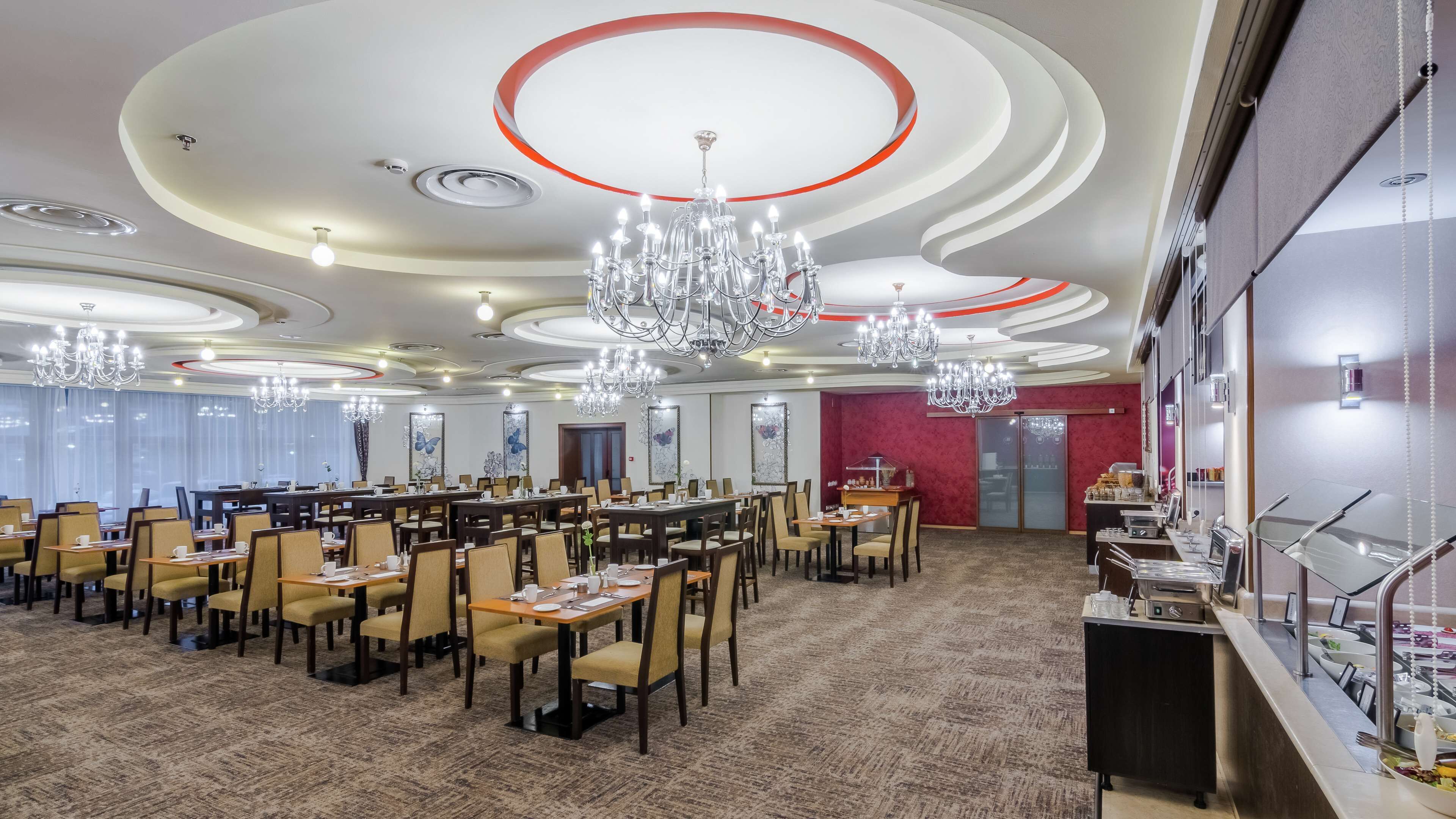 A spacious dining area with multiple tables and chairs, chandeliers, and a buffet counter.