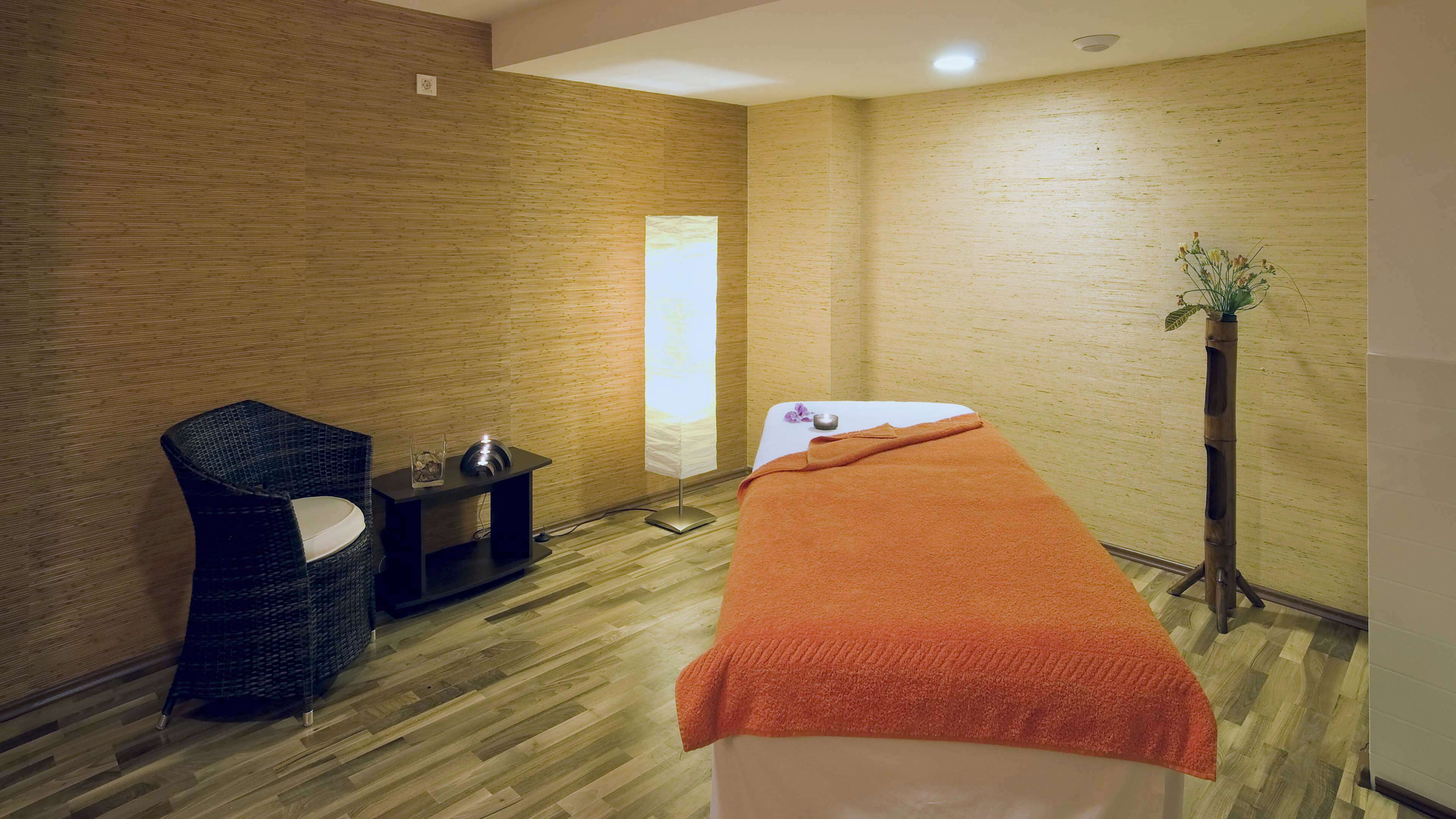 A room with a massage table, a small side table with a candle, a chair, and a tall plant.