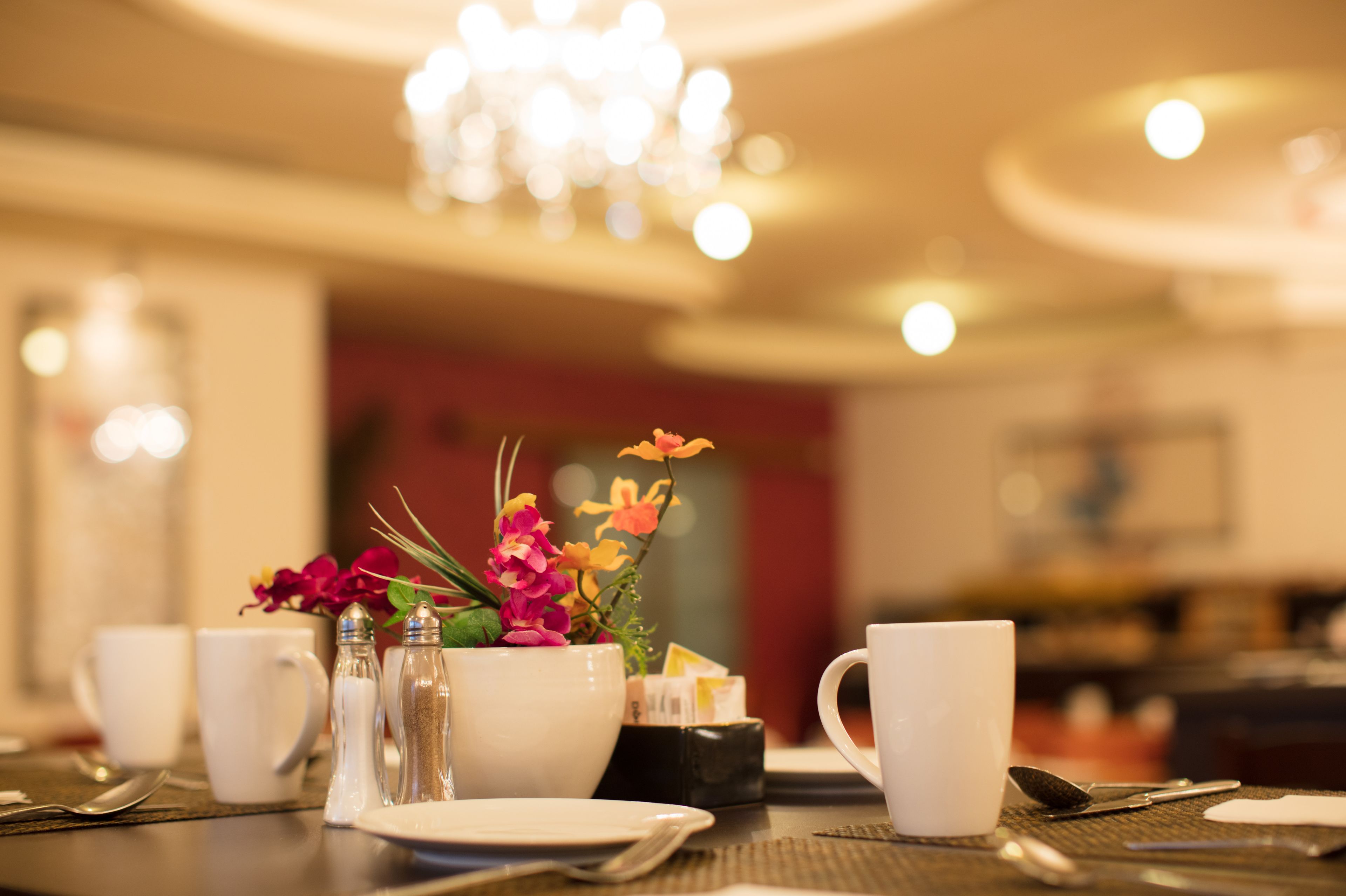 Una mesa de desayuno dispuesta con tazas, platos y cubiertos blancos, un salero y pimentero, y un centro de mesa floral.