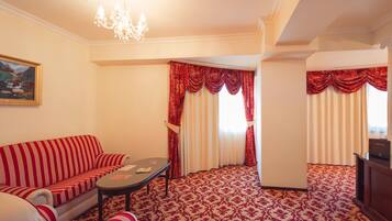A hotel room with a red and white striped sofa, a small table, a painting on the wall, a chandelier, red curtains, a television, and a patterned carpet.