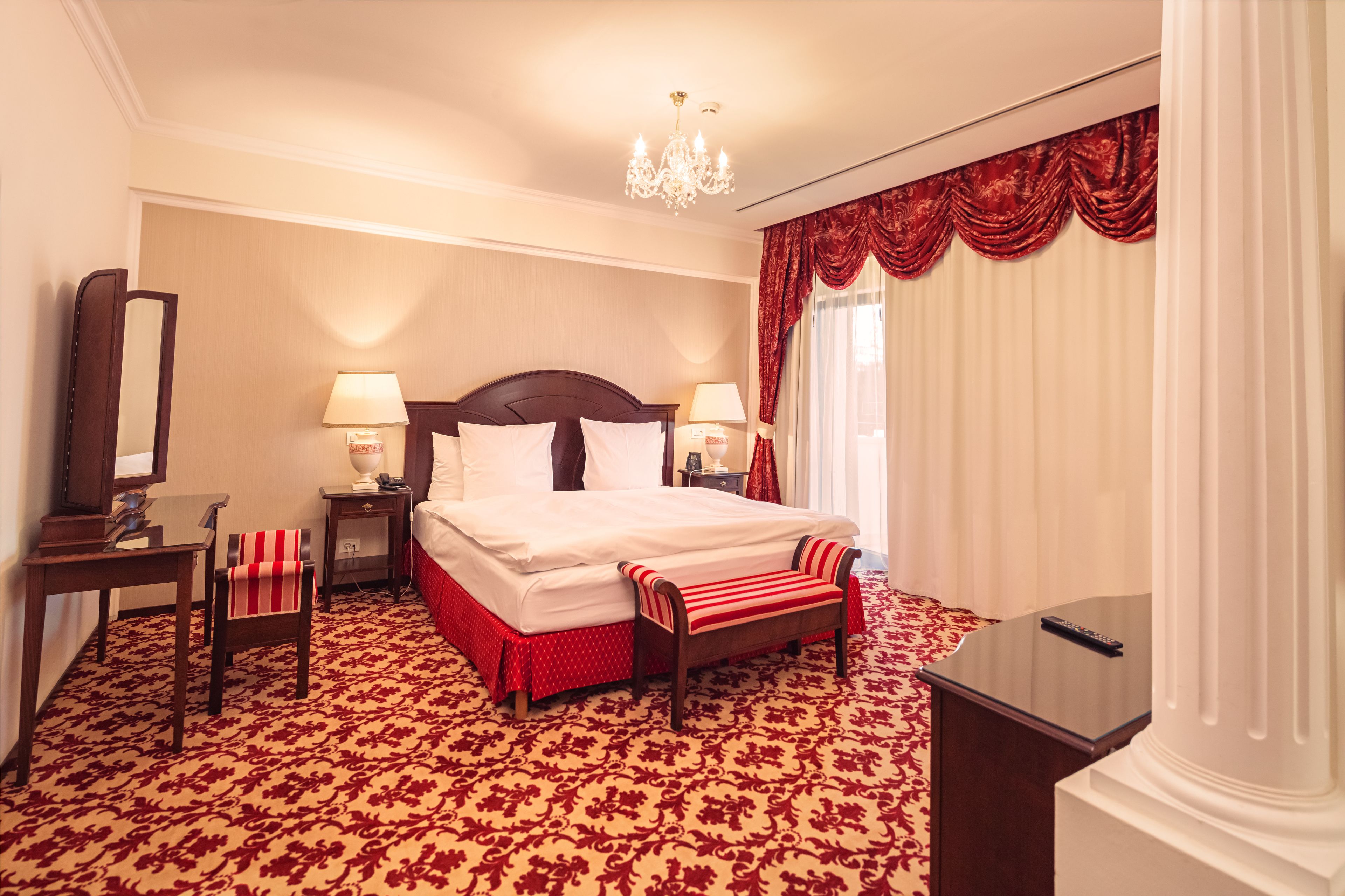 Una habitación de hotel con una cama grande, dos mesitas de noche con lámparas, un escritorio, una silla, un banco y una araña.