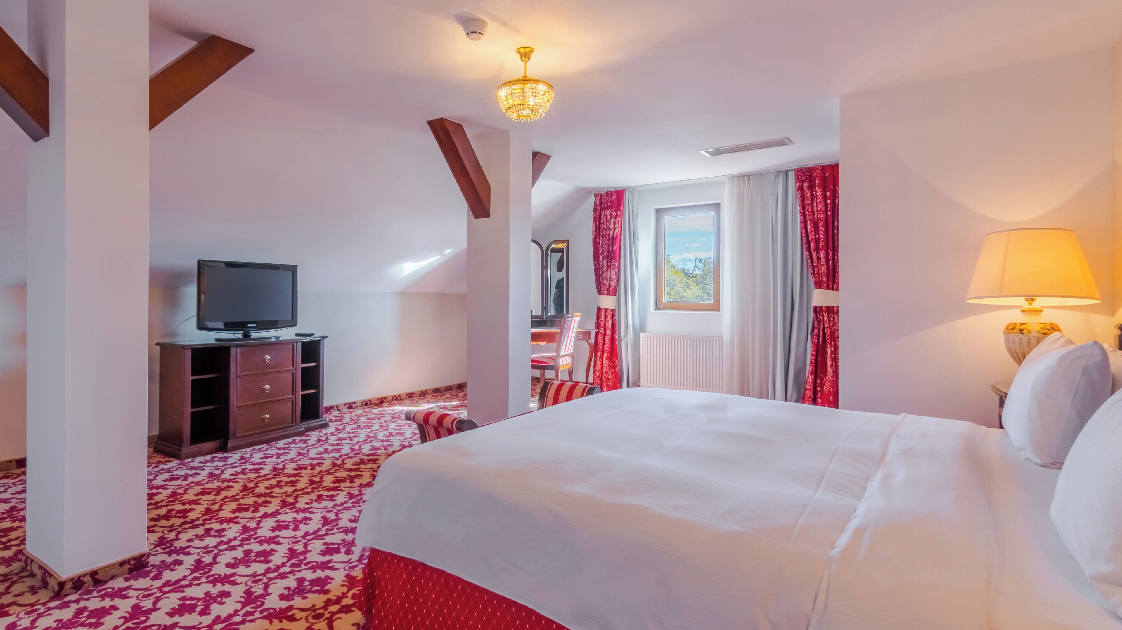 Ein Hotelzimmer mit einem großen Bett, einem Fernseher auf einer Holzkommode, einem Schreibtisch mit Stuhl und einem Fenster mit Vorhängen.