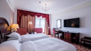 A hotel room with two beds, a desk, a television, and a chandelier.