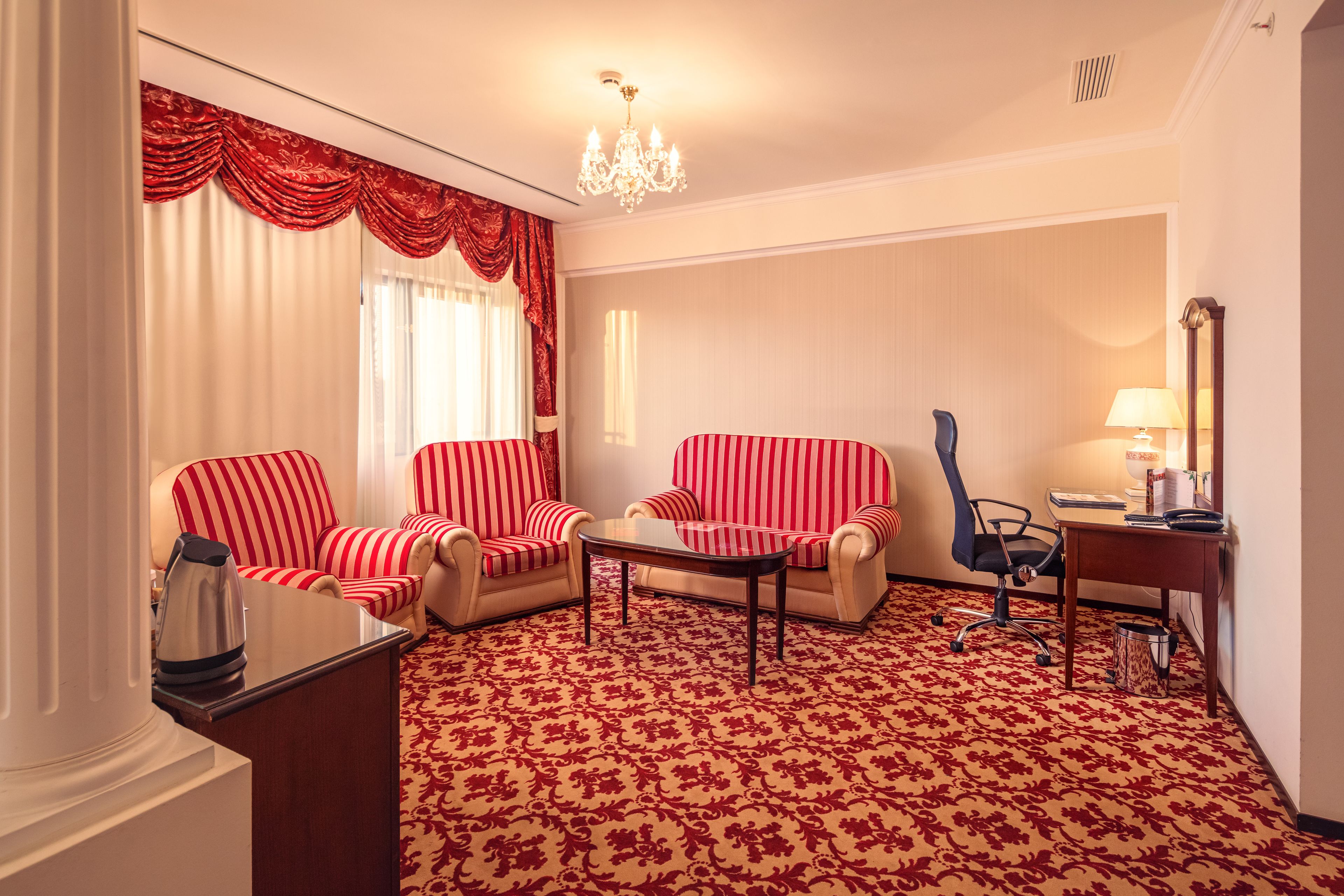 A hotel room with two striped armchairs, a small table, a desk with a lamp, and a patterned carpet.
