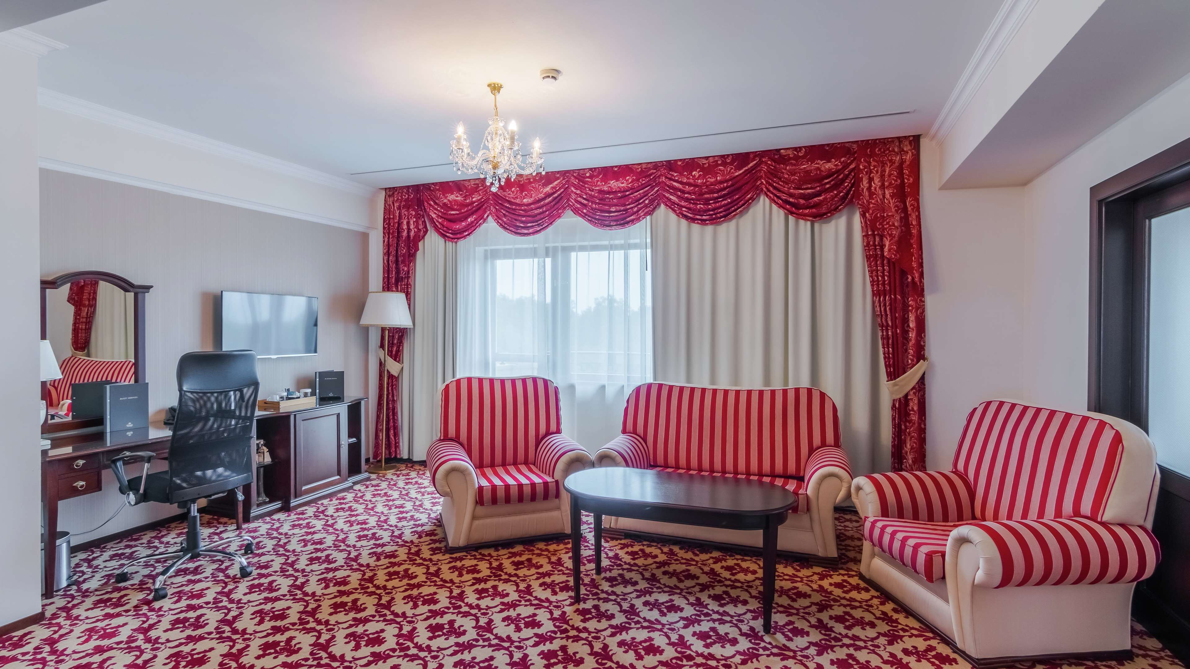 A hotel room with a large window, a chandelier, two striped sofas, a desk with a computer, and a patterned carpet.