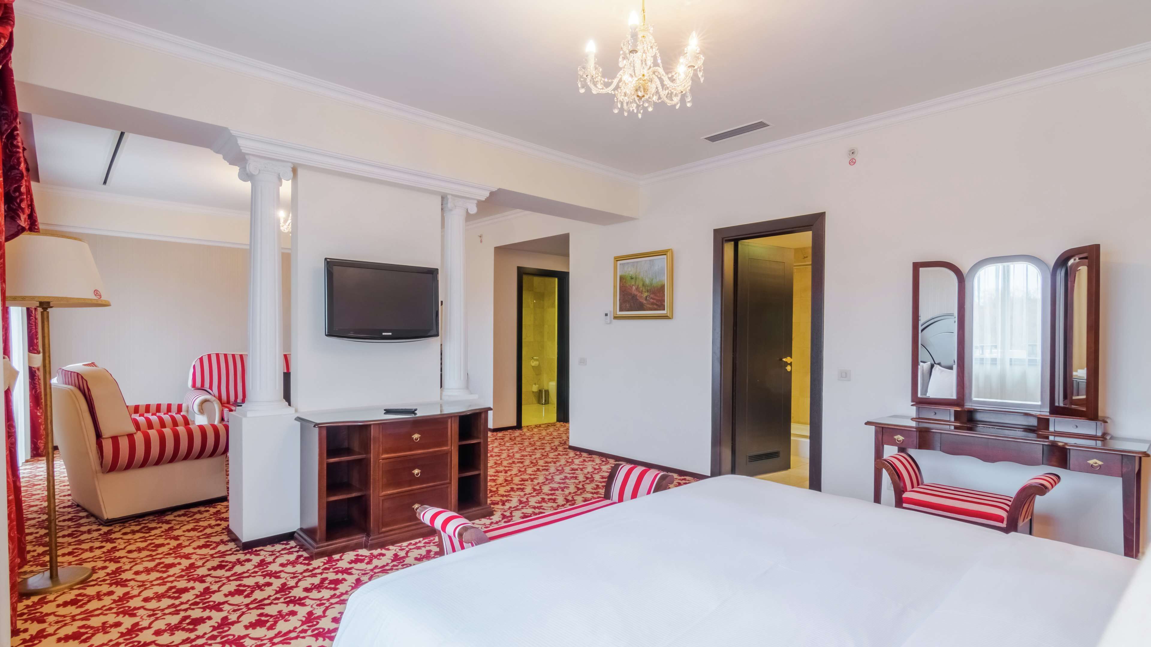 A hotel room with a large bed, a TV mounted on the wall, a desk with a chair, a mirror, and a door leading to another room.