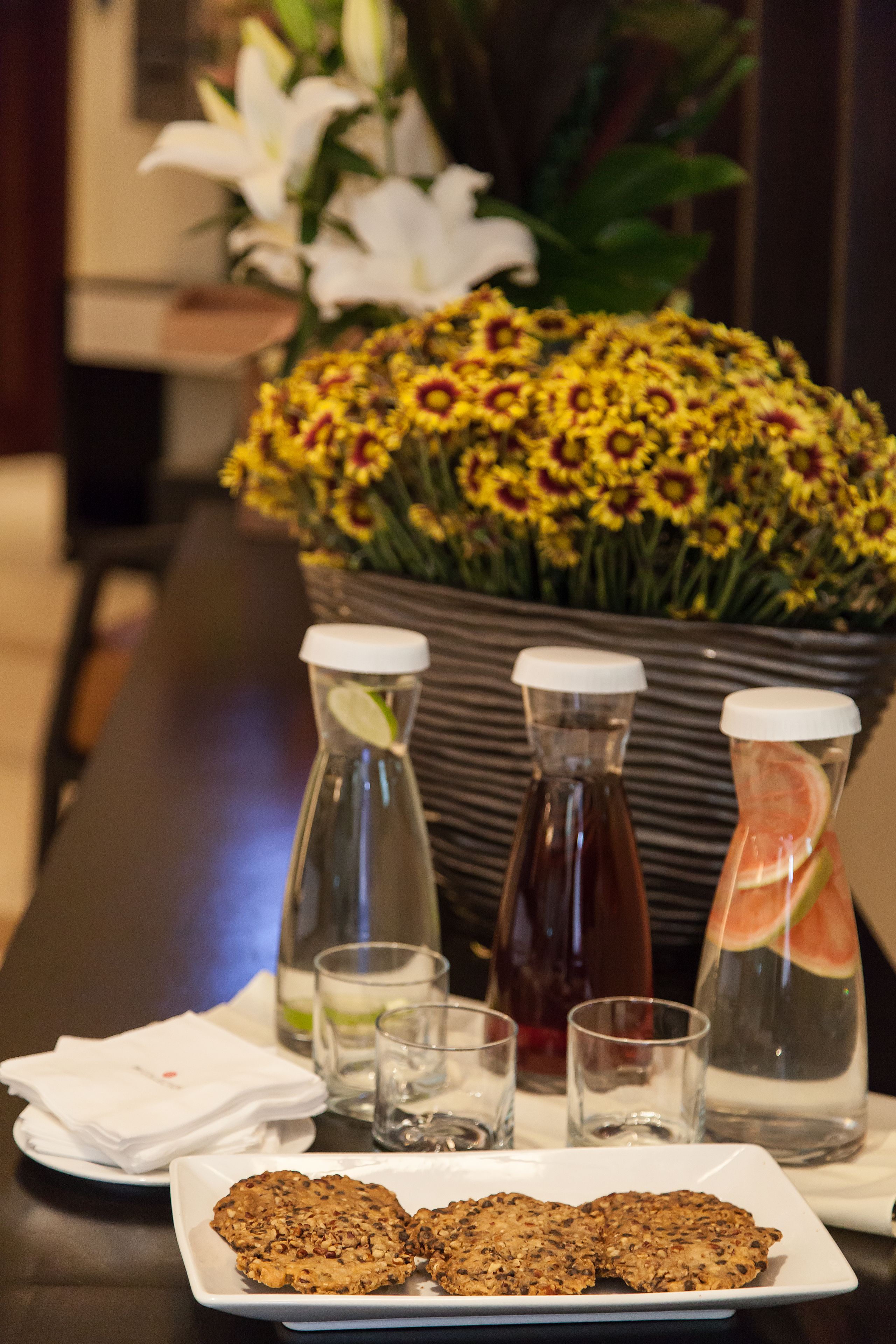 A table with a plate of cookies, two bottles of water, and a pitcher with a slice of watermelon. There is a vase of yellow flowers in the background.