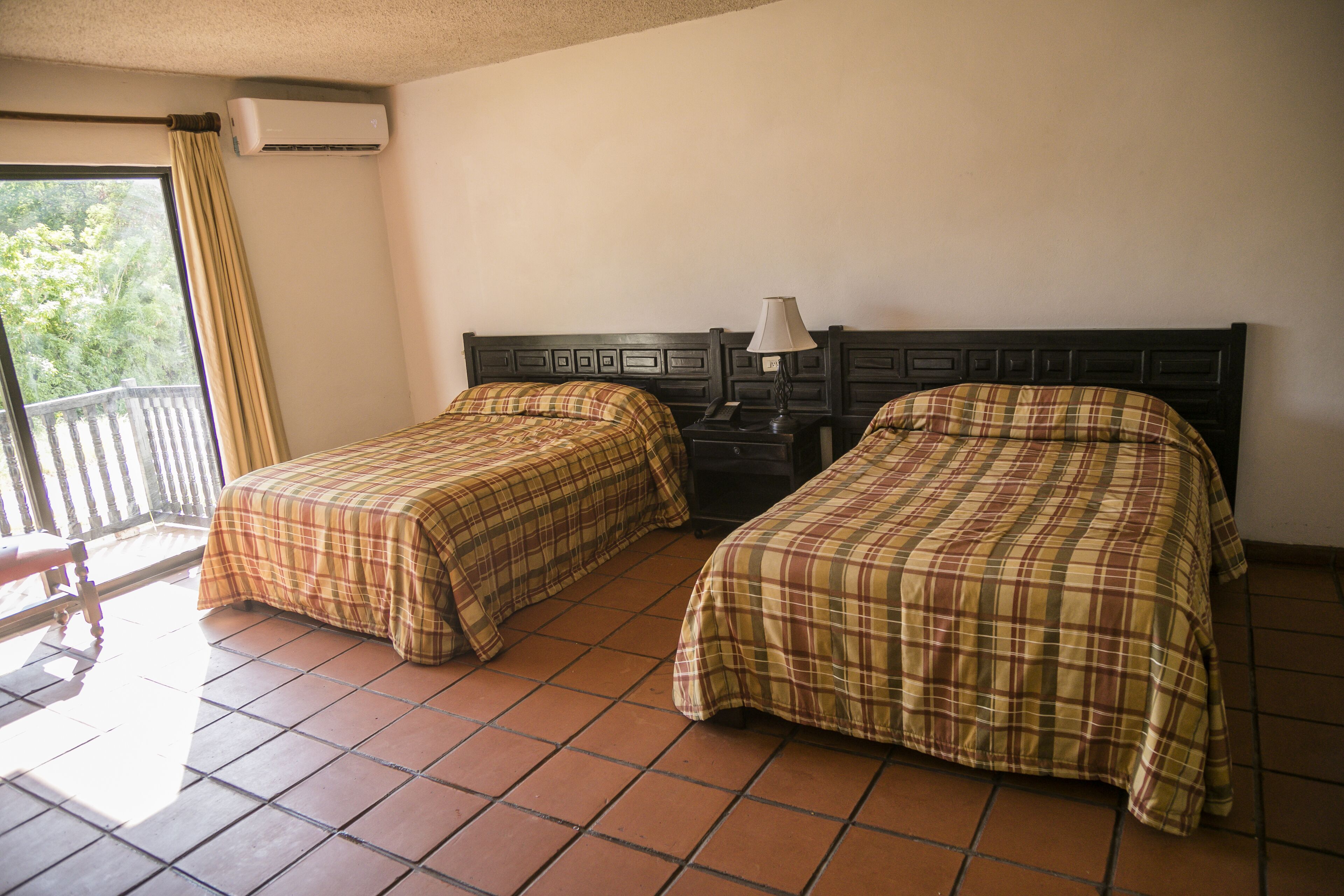 Una habitación con dos camas, una ventana con cortinas, aire acondicionado y una mesita de noche con lámpara.