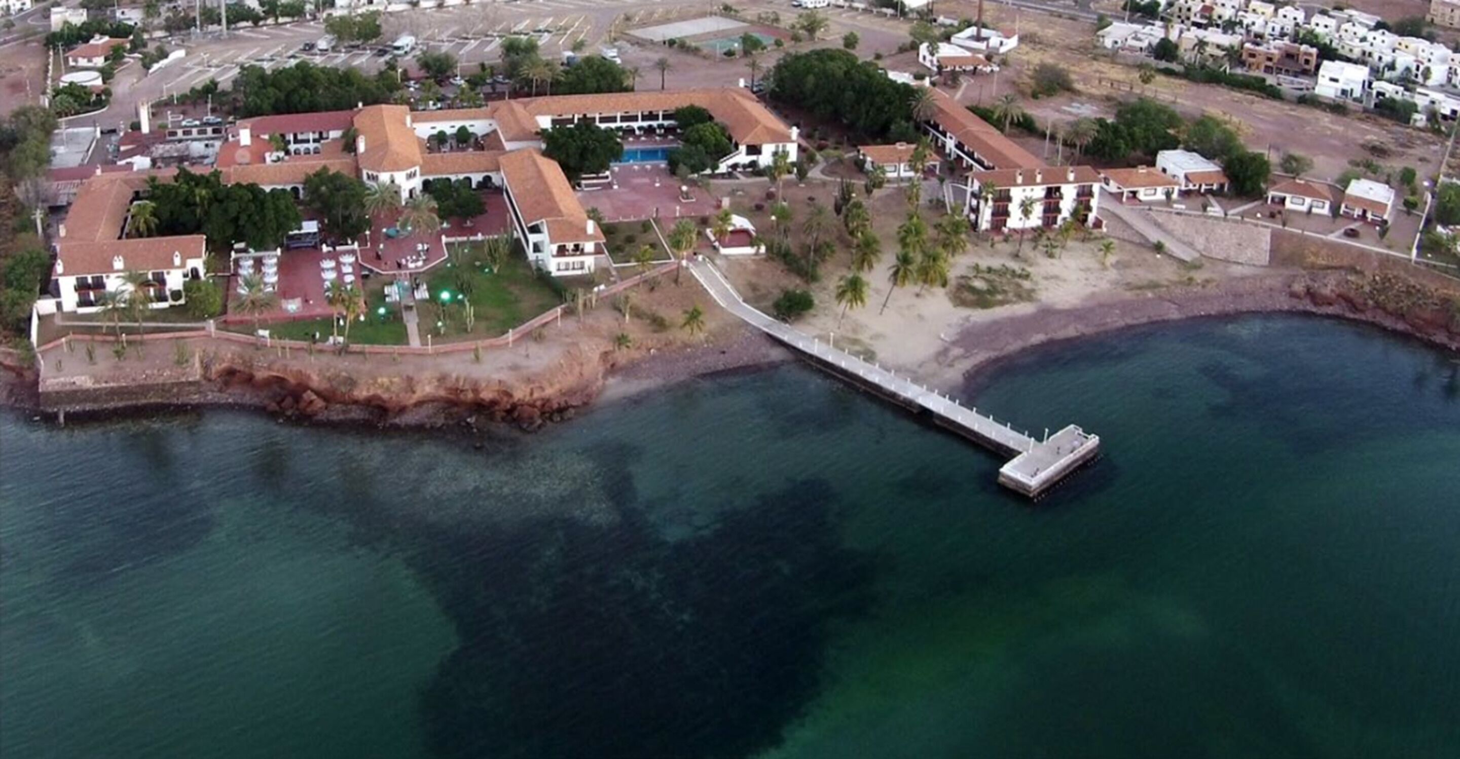 Vista aérea de una zona costera con un muelle que se adentra en el agua, rodeada de edificios residenciales y vegetación.