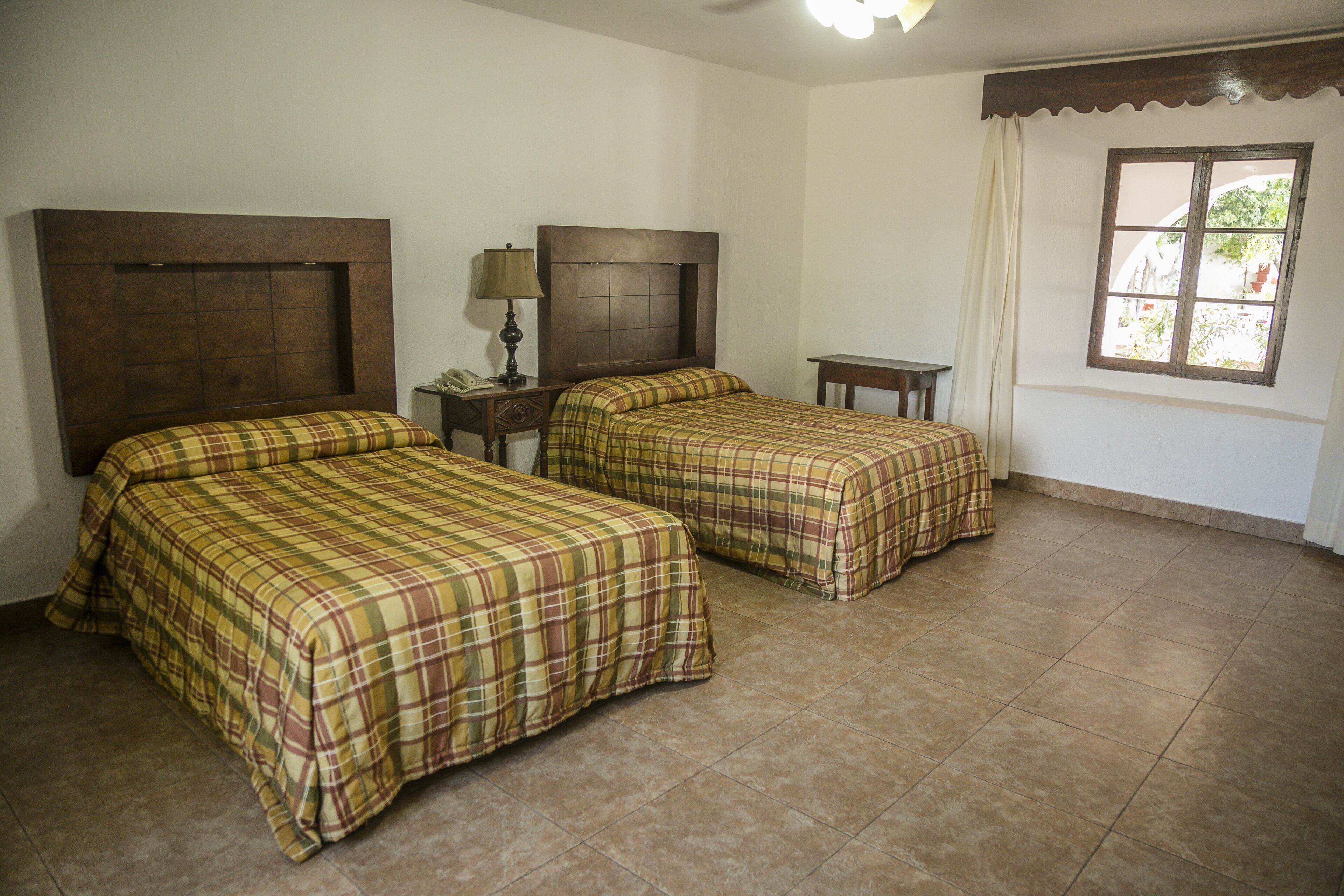 Una habitación con dos camas, una mesita de noche, una lámpara y un ventanal con cortinas.