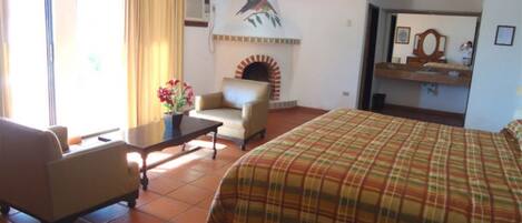 Habitación de hotel con cama, sofá, mesa con flores, chimenea y baño visible a través de una puerta abierta.