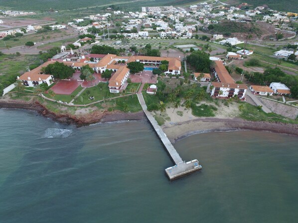 Vista aérea de un pueblo costero con un muelle que se adentra en el mar, rodeado de edificaciones residenciales y zonas verdes.