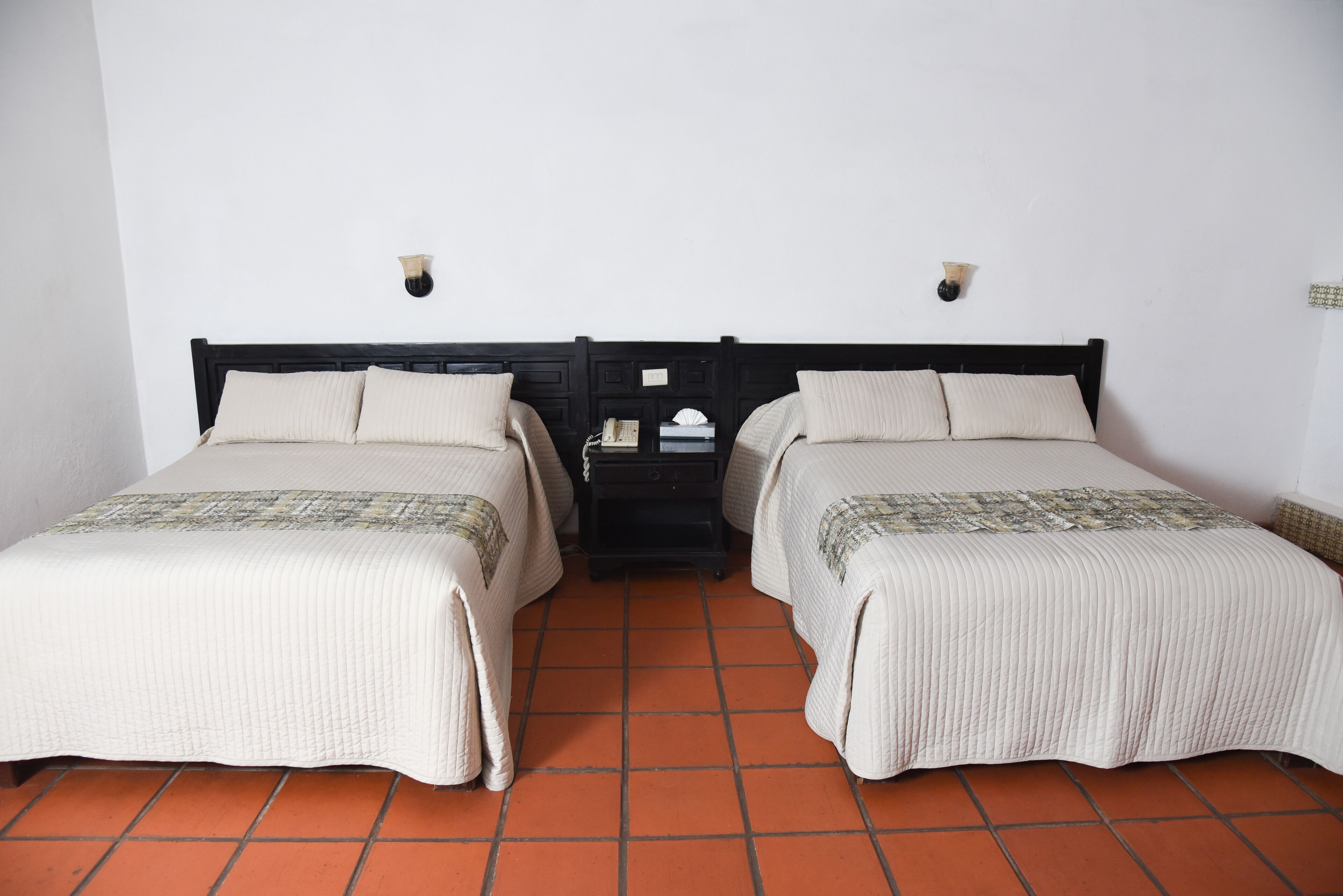 Una habitación con dos camas, cada una con cabecera y almohadas, una mesita de noche con lámpara y suelo de baldosas.