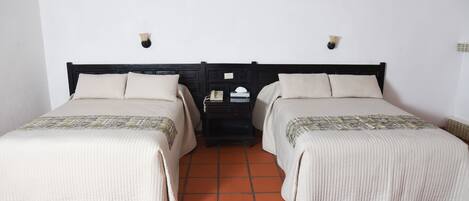 Una habitación con dos camas, cada una con cabecera y almohadas, una mesita de noche con lámpara y suelo de baldosas.