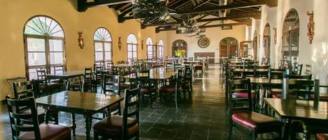 A spacious dining hall with high ceilings, large windows, and multiple dining tables and chairs.