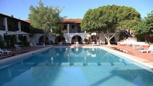 Una piscina con sillones y sombrillas, rodeada por un edificio con arcos y árboles.